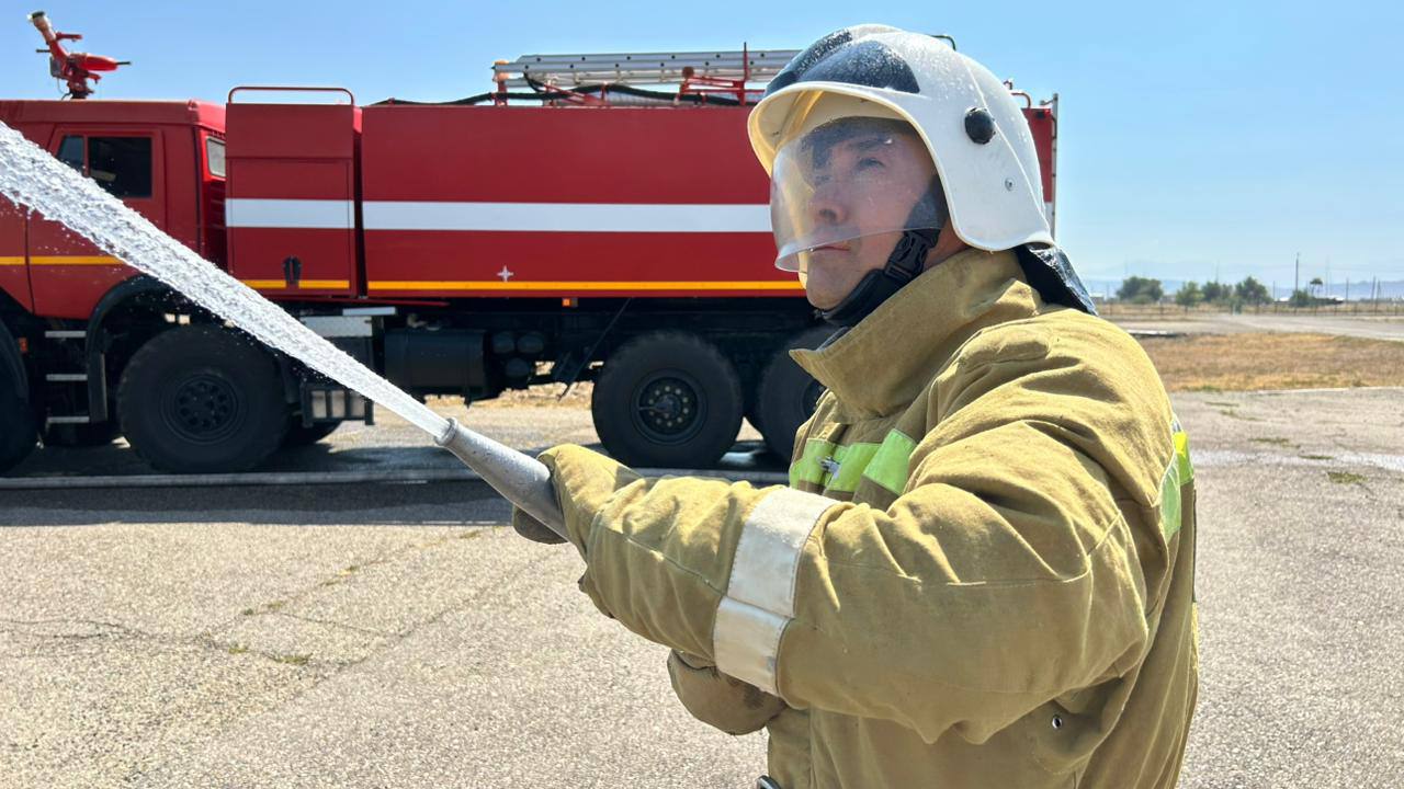 Өрт сөндіруші арнайы киімде су шашатын шлангті ұстап тұр, артқы жағында қызыл өрт сөндіру көлігі.