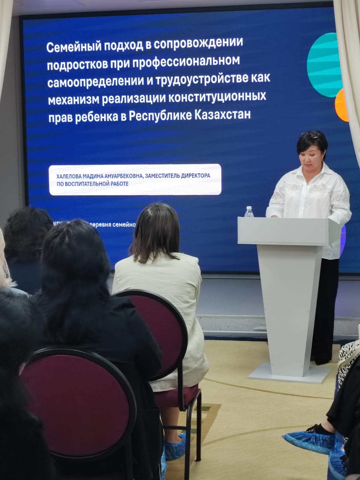 Сөйлеуші әйел экрандағы презентация бойынша баяндама жасап тұр, ал алдында тыңдаушылар отыр.