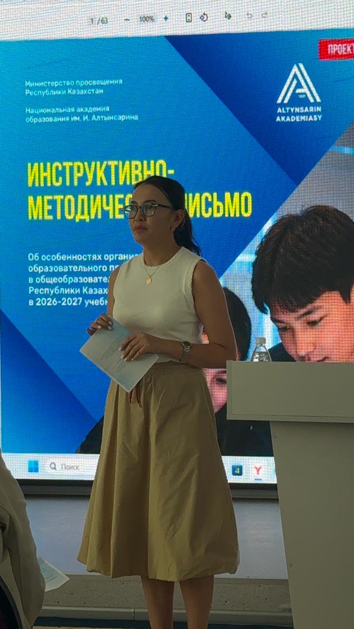 Презентация алдында тұрған, қолында қағазы бар әйел адам. Фонда «Алтынсарин академиясының» нұсқаулық-әдістемелік хаты бейнеленген.