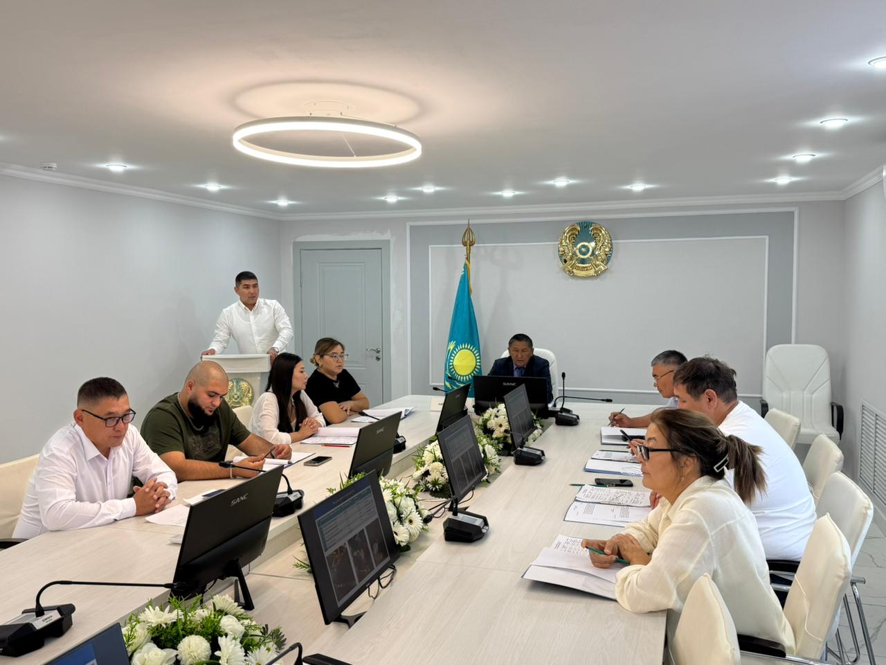 Жиналыс залындағы ресми кеңес: үстел басында отырған қатысушылар және баяндама жасап тұрған адам.