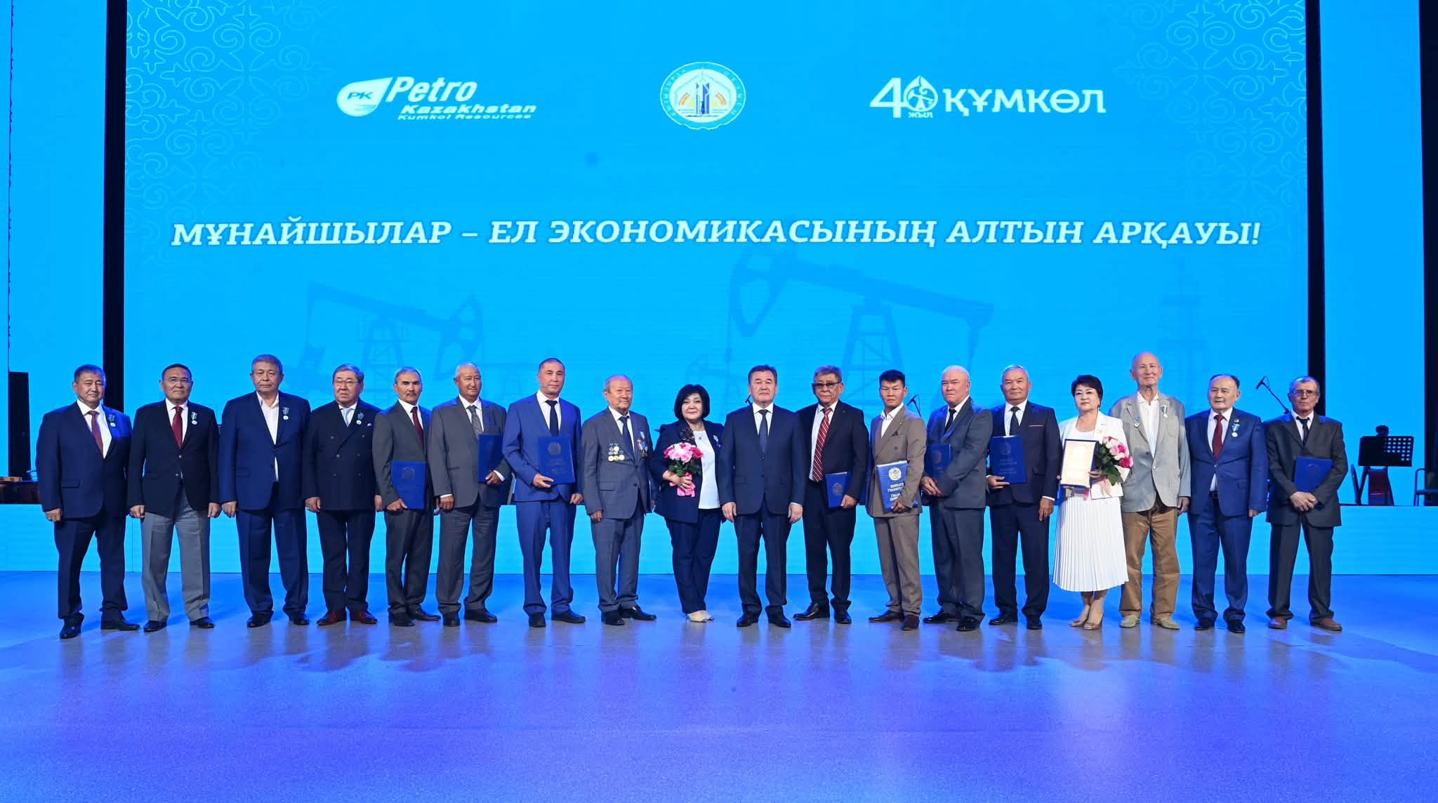 Көк фонды сахнада «Мұнайшылар – ел экономикасының алтын арқауы!» жазуымен тұрған сыйлықарт алған адамдар тобы.