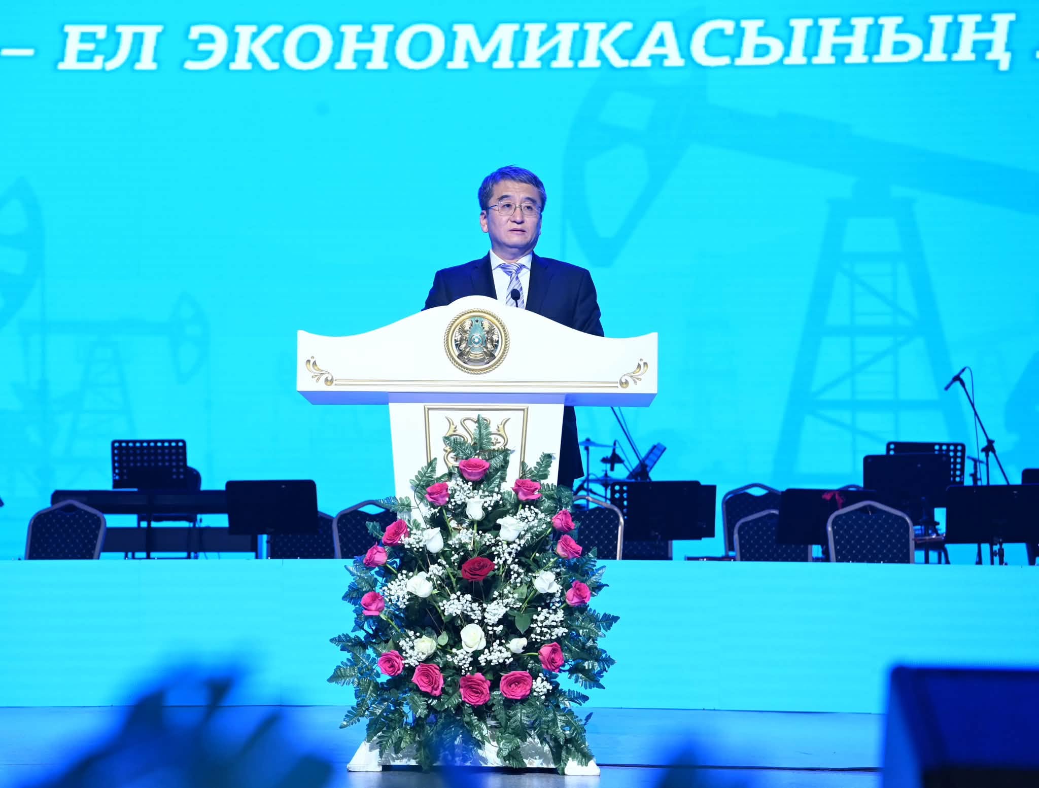 Көк фонды экранның алдында, гүлдермен безендірілген трибунада сөйлеп тұрған костюм киген ер адам.