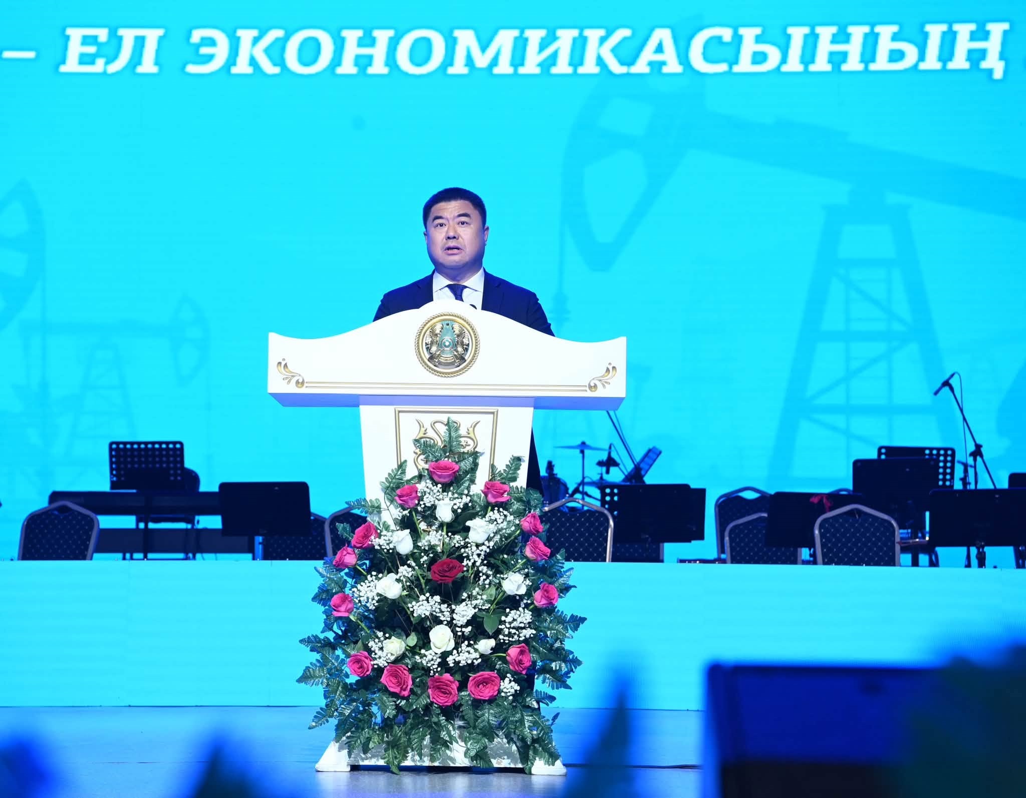 Көк түсті фон алдындағы трибунада сөйлеп тұрған ер адам. Трибуна гүлдермен безендірілген.