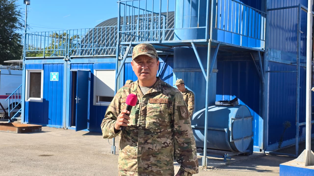 Камуфляж киген әскери адам микрофонмен көк түсті контейнерлердің алдында сөйлеп тұр.