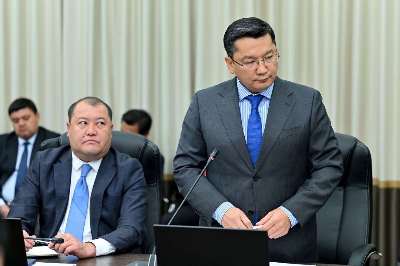Костюм киген ер адам жиналыста сөйлеп тұр, ал жанында басқа әріптестері отыр.