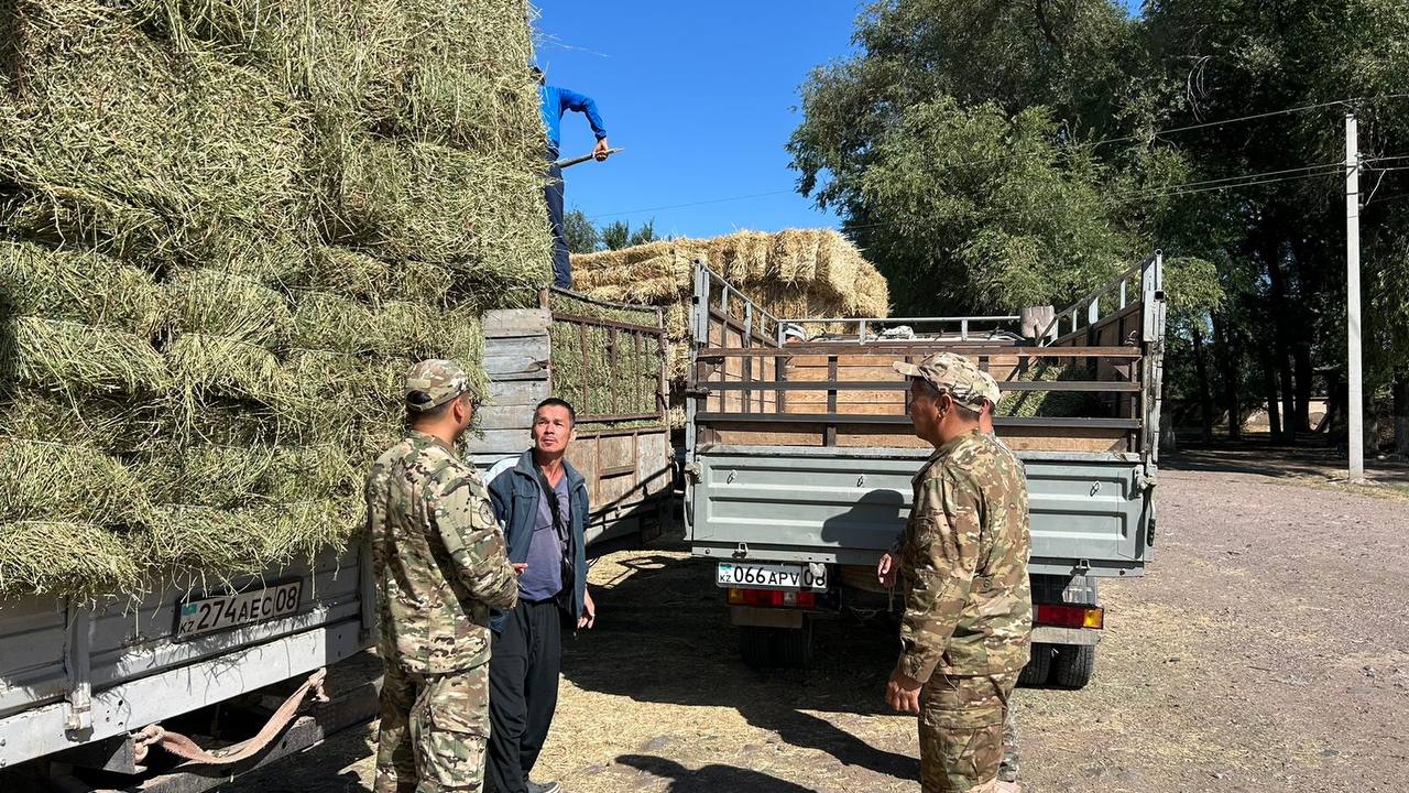 Шөп тиелген жүк көліктерінің жанында тұрған әскерилер мен жұмысшы.