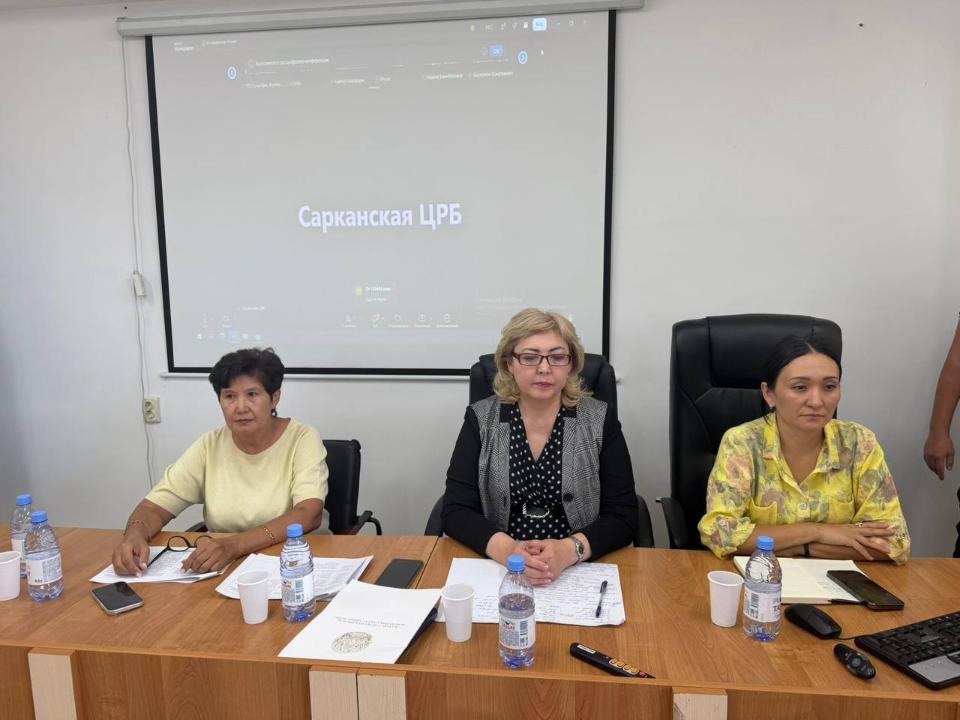 Үстел басында отырған үш әйел және артқы жақтағы «Сарканская ЦРБ» жазуы бар проектор экраны.