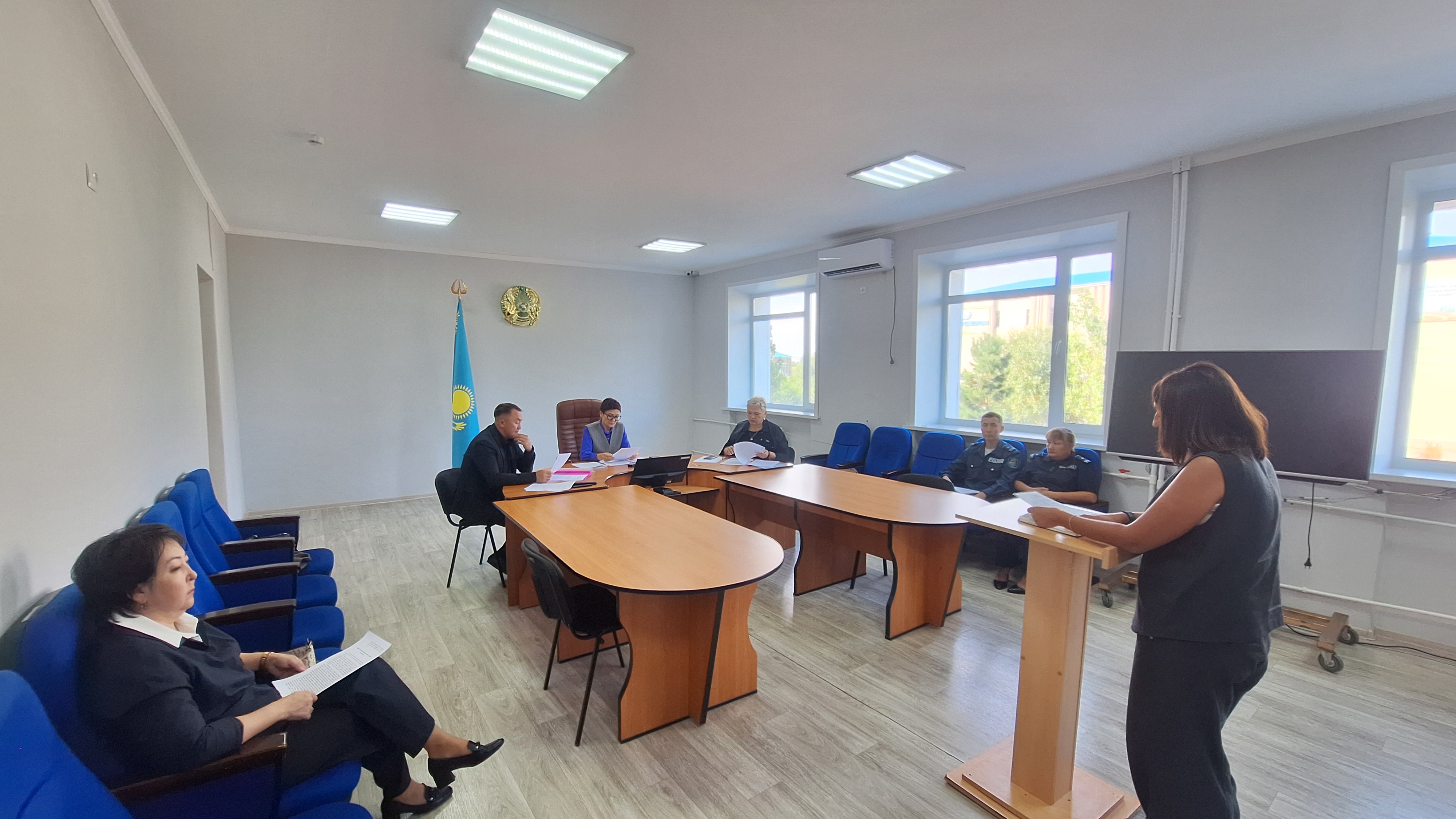 Жылынамадағы мәжіліс: трибунада сөйлеп тұрған әйел және үстел басында отырған қатысушылар.