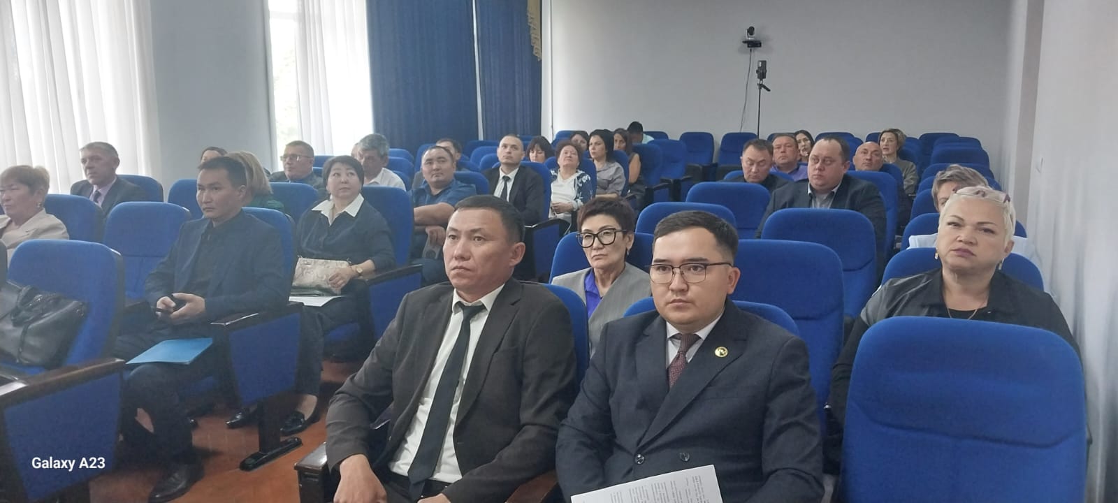 Көк орындықтарда отырған костюм киген адамдар тобы.