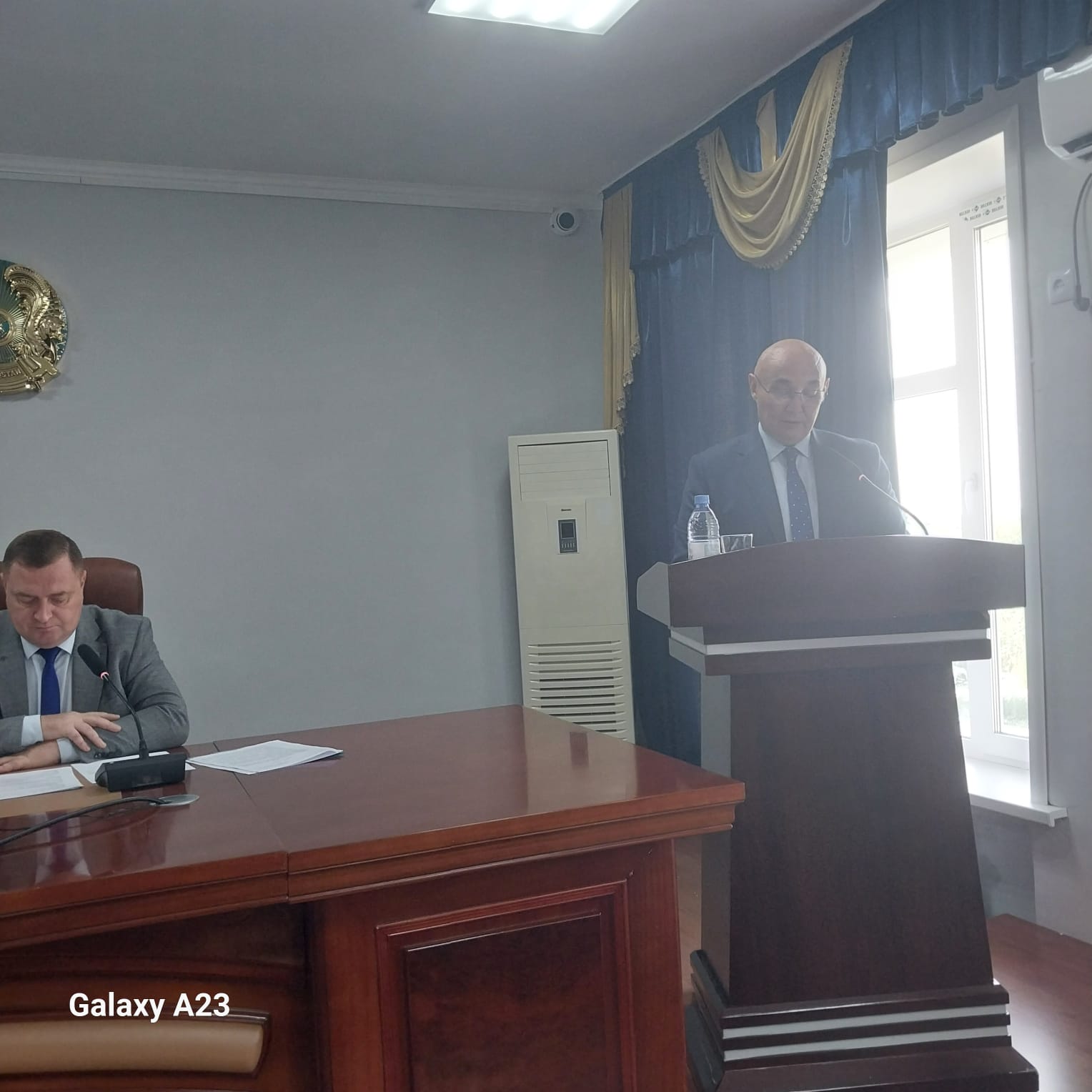 Трибунада сөйлеп тұрған ер адам және үстелде отырған әріптесі.