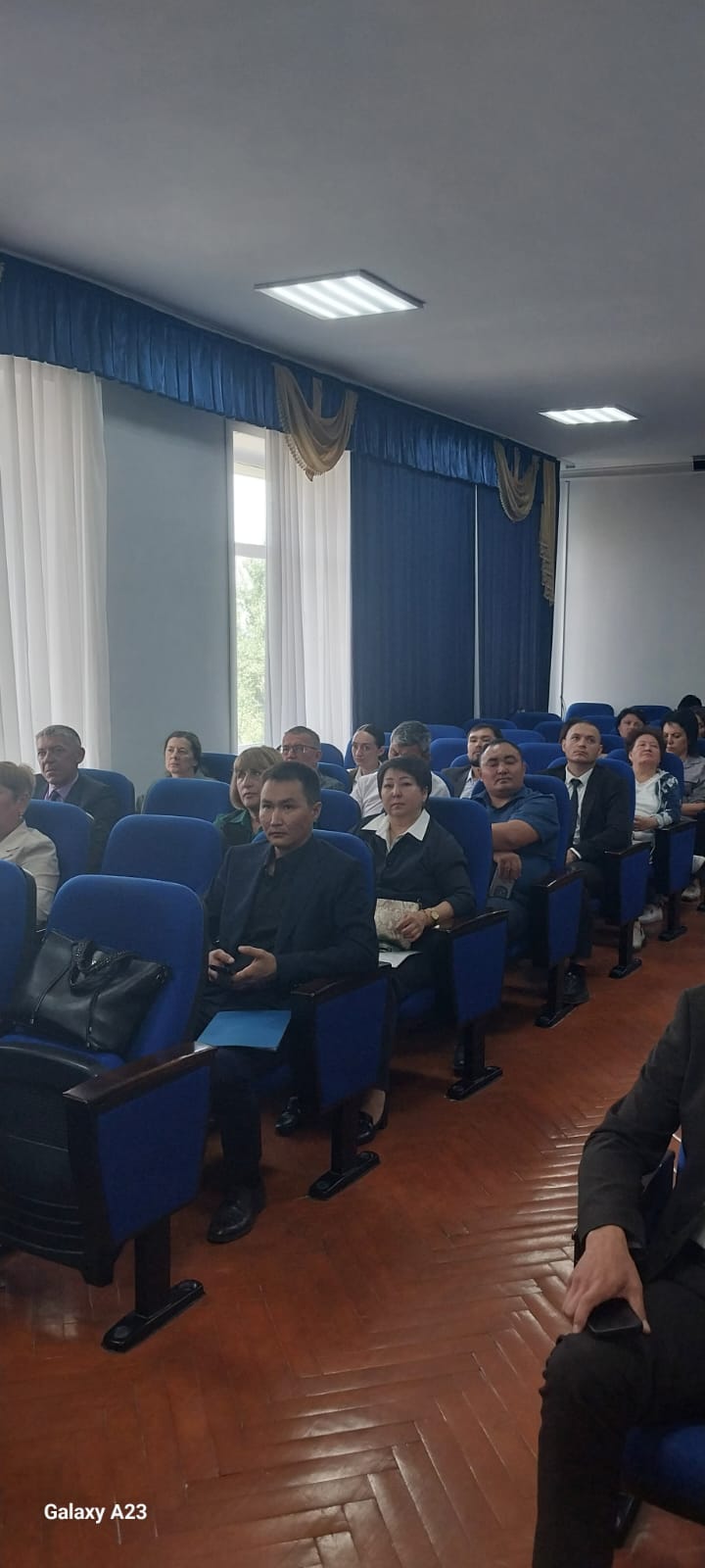 Көк орындықтарда отырған адамдар тобы жиналыста немесе конференцияда тыңдап отыр.