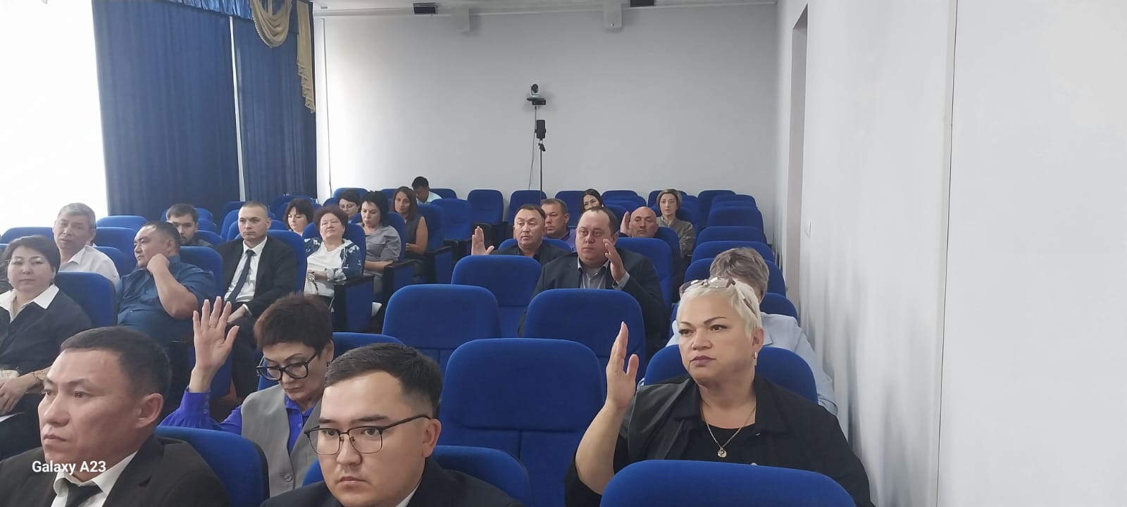 Көк орындықтарда отырған адамдардың қолын көтеріп дауыс беруі.