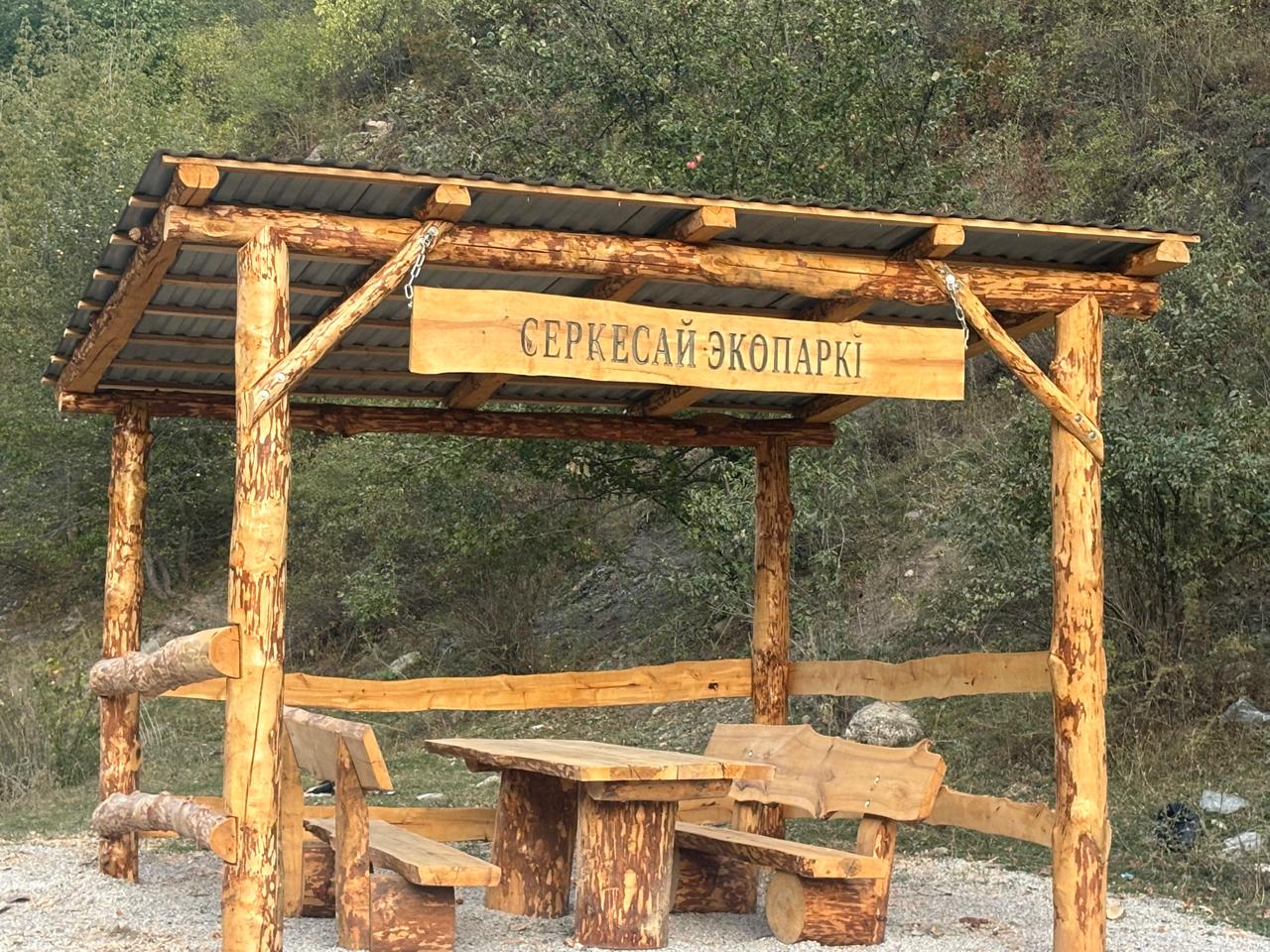 Деревянная беседка с навесом, столом и скамьями. Сверху вывеска «СЕРКЕСАЙ ЭКОПАРКІ» на фоне зеленого леса.