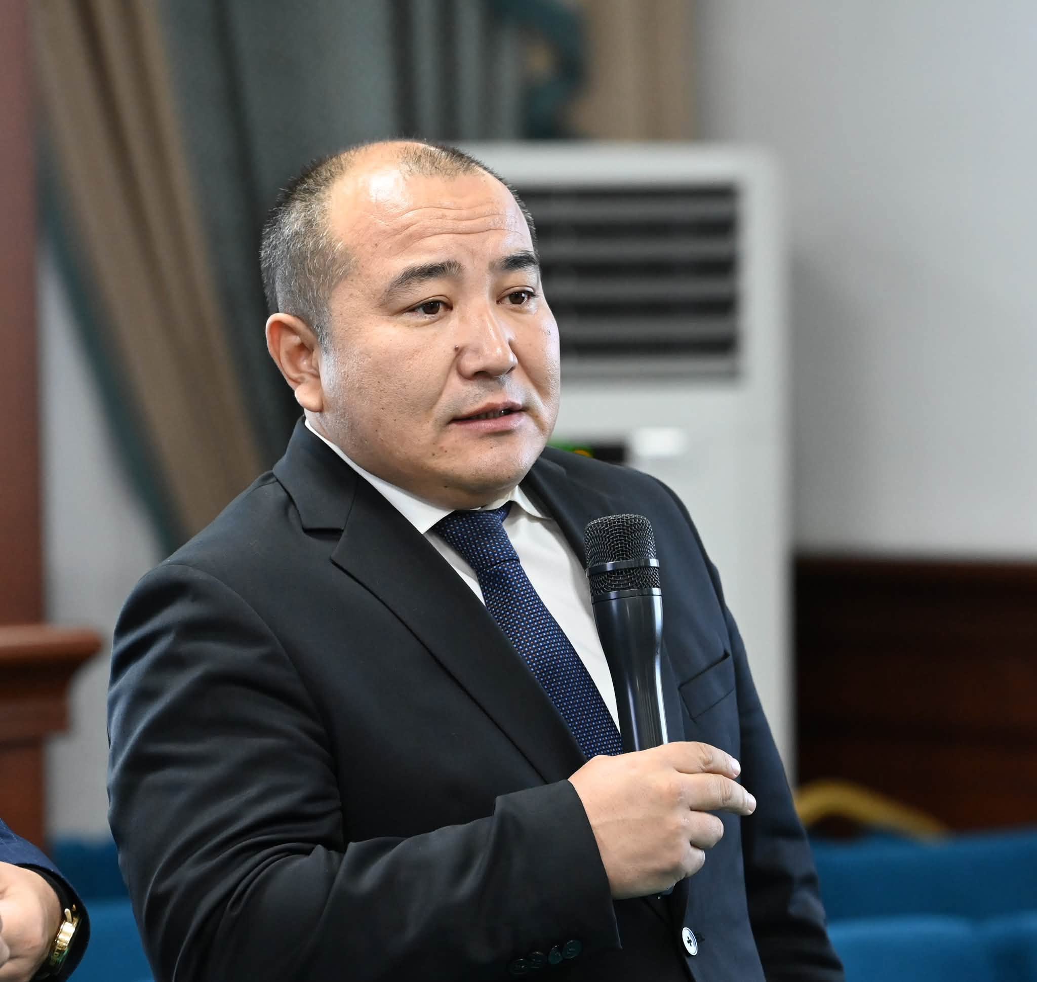 Қара костюм киген, қолына микрофон ұстаған ер адамның портреті.