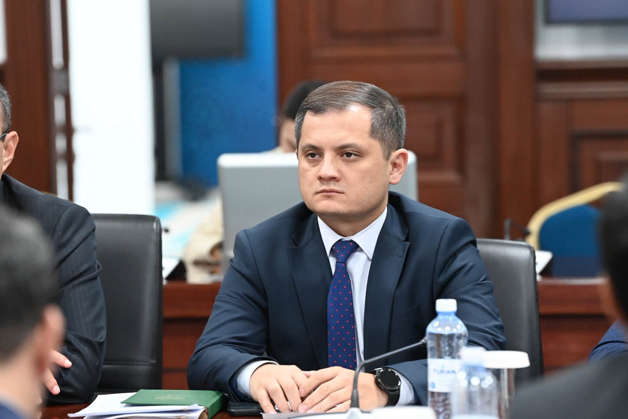 Көк костюм мен нүктелі галстук киген ер адам жиналыста отыр.
