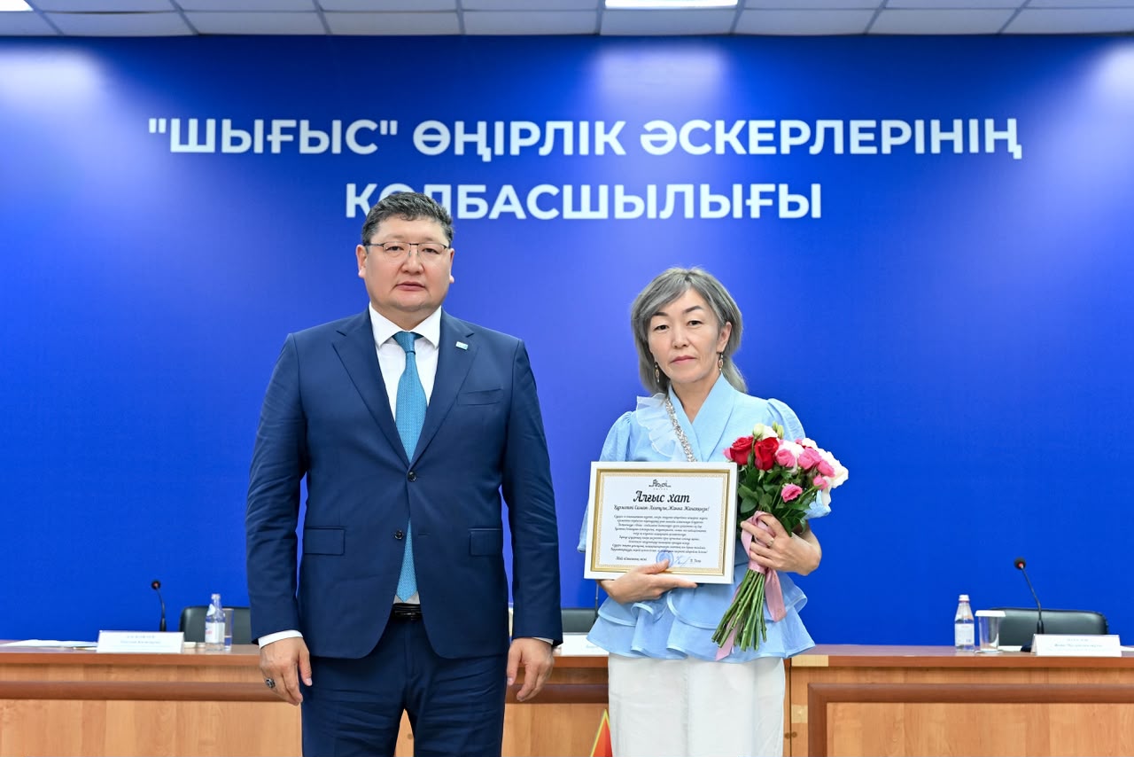 «Шығыс» өңірлік әскерлерінің қолбасшылығы алдында грамота және гүл ұстаған әйел адам.