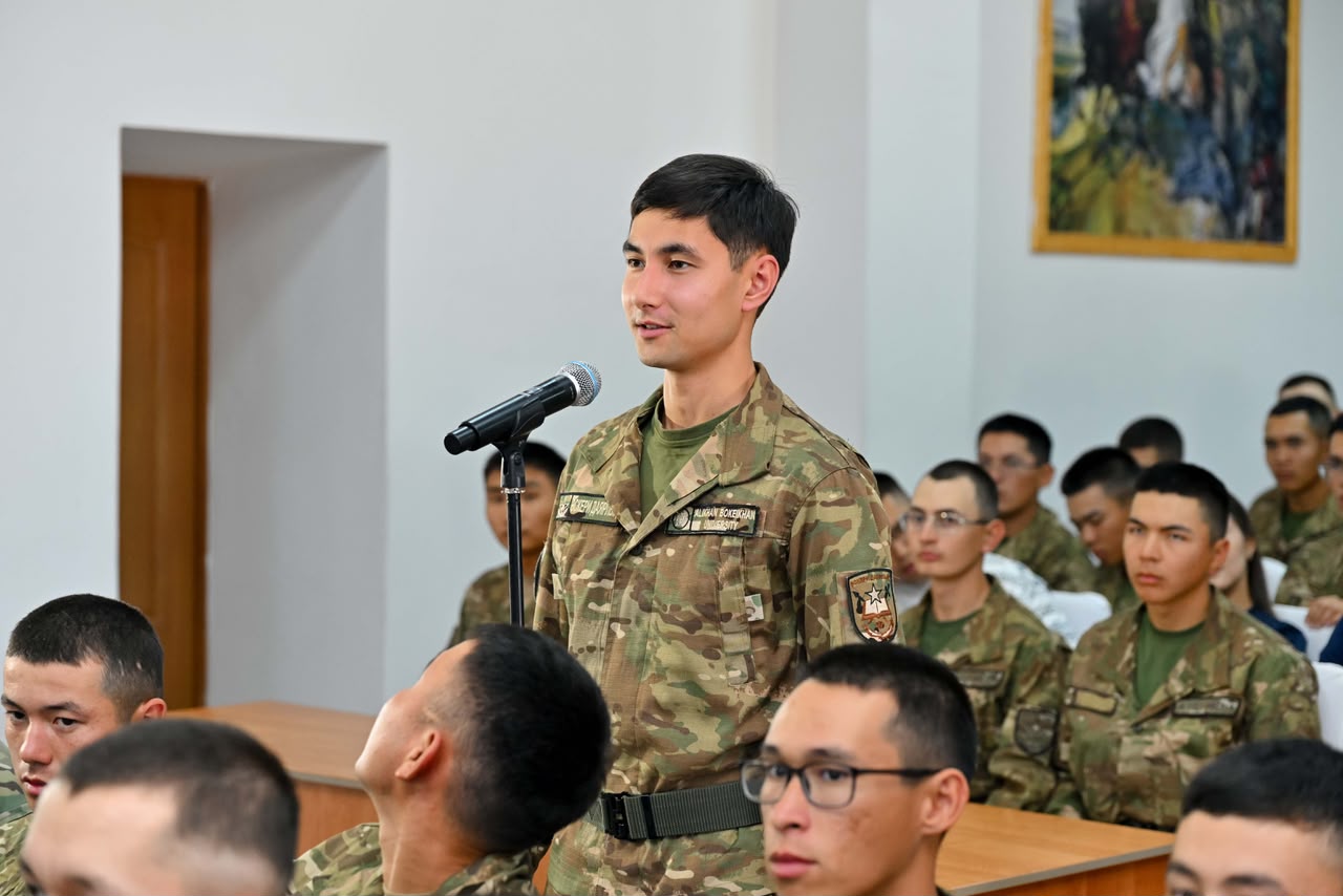 Микрофон алдында сөйлеп тұрған әскери формадағы жас сарбаз және оны тыңдап отырған әріптестері.