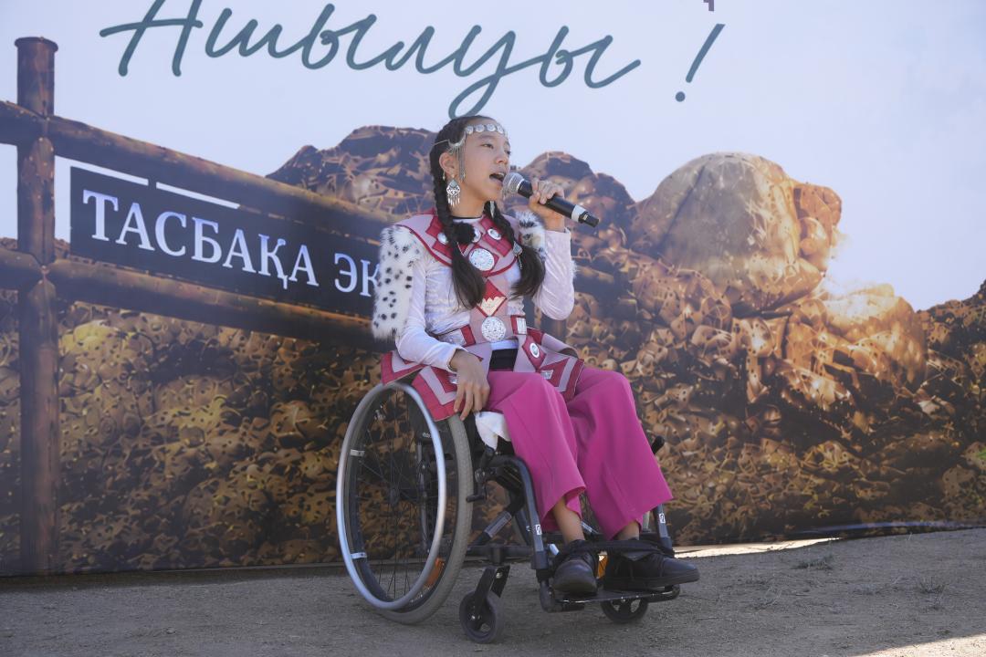 Ұлттық киім киген, арбада отырған қыз микрофонмен сөйлеп тұр.