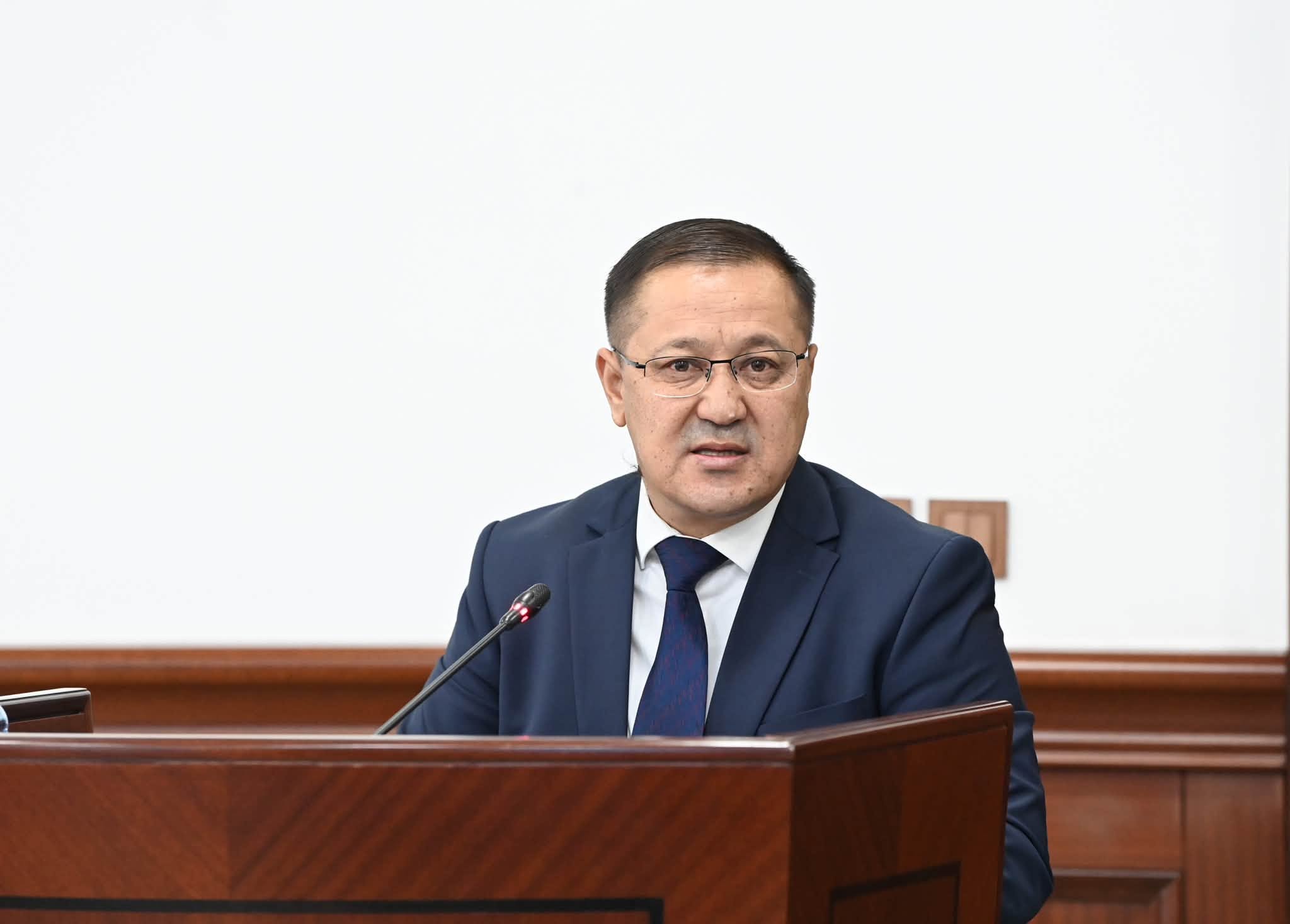 Көзілдірік пен костюм киген ер адам трибунада сөйлеп тұр.
