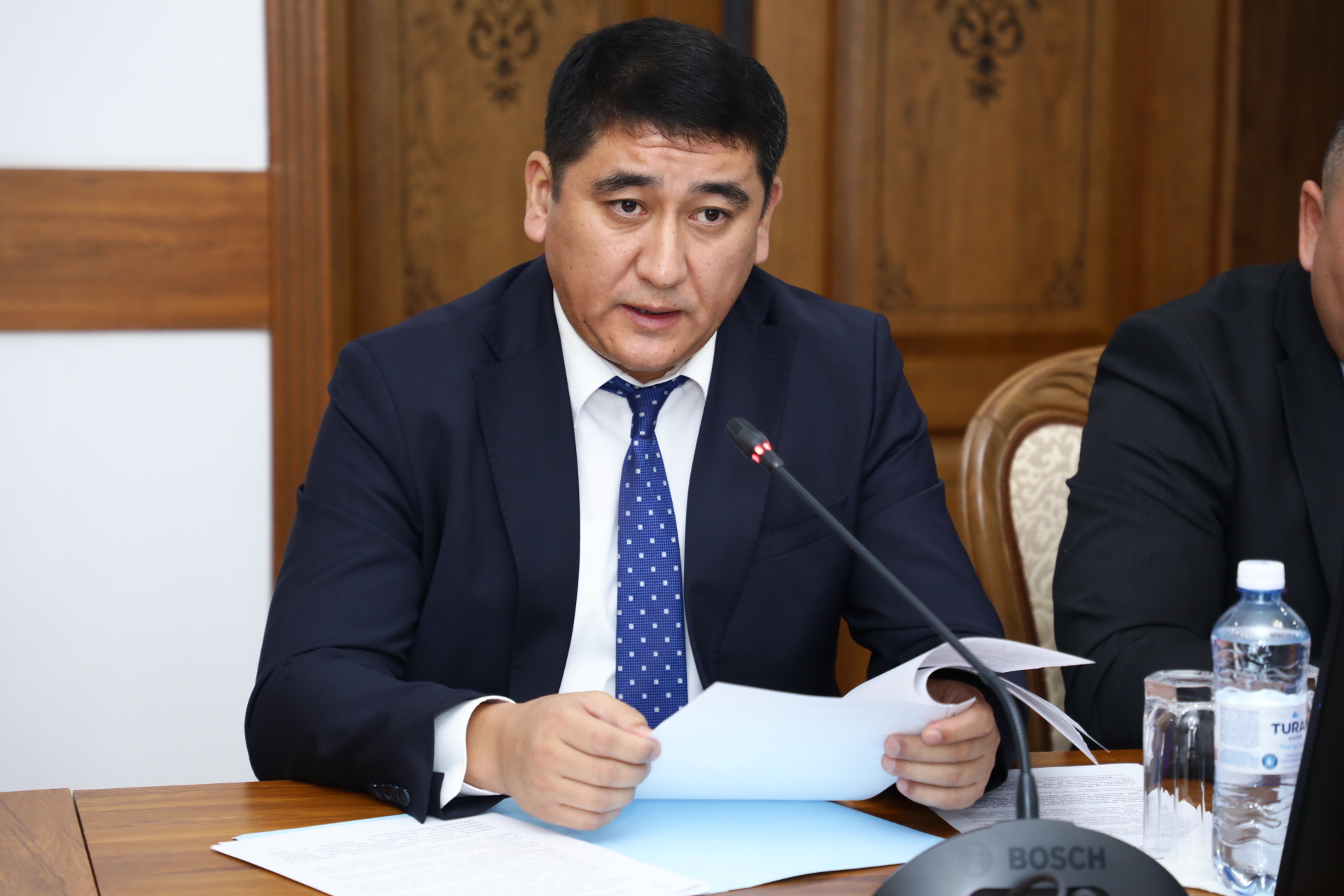Көк костюм киген ер адам микрофонның алдында қағаздарды оқып отыр.