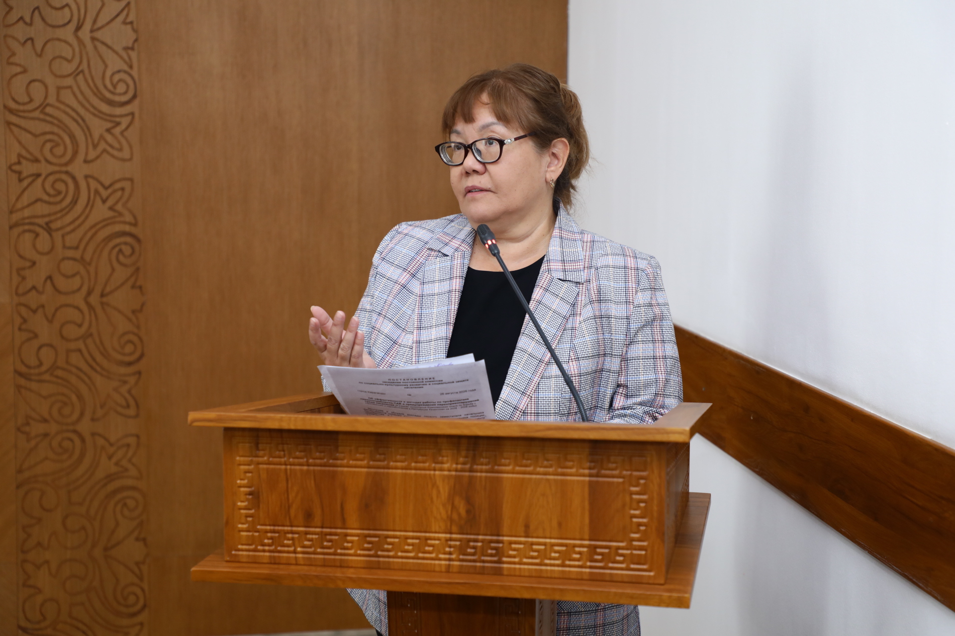 Көзілдірік киген әйел адам трибунада тұрып, қағазға қарап сөйлеп жатыр.