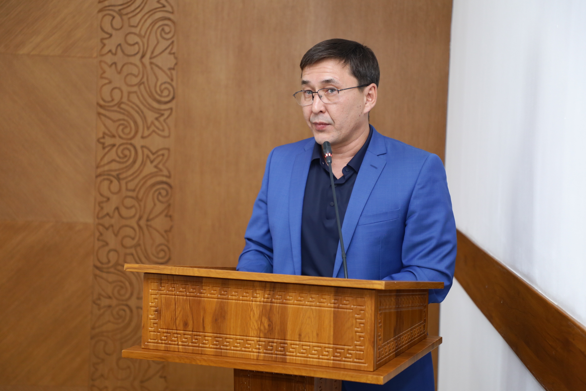 Көк костюм киген, көзілдірікті ер адам ағаш трибунада сөйлеп тұр.