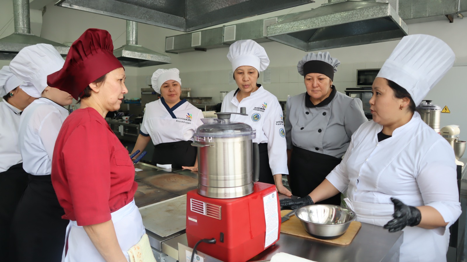 Группа студентов в профессиональной форме стоят на кухне вокруг кухонного оборудования.