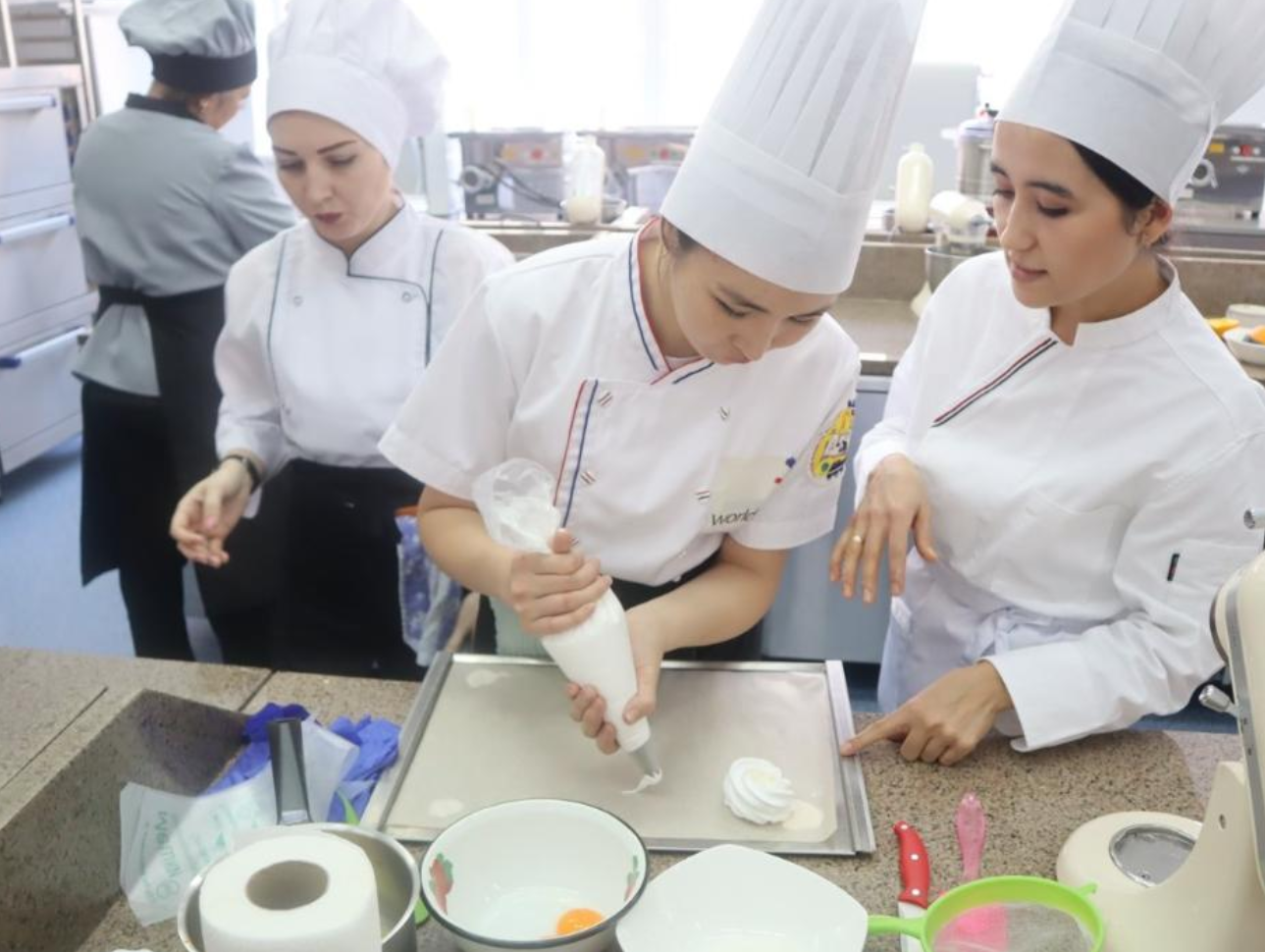 Ақ киімді және повар қалпағын киген үш кондитердің тәттілер пісіру процесі.
