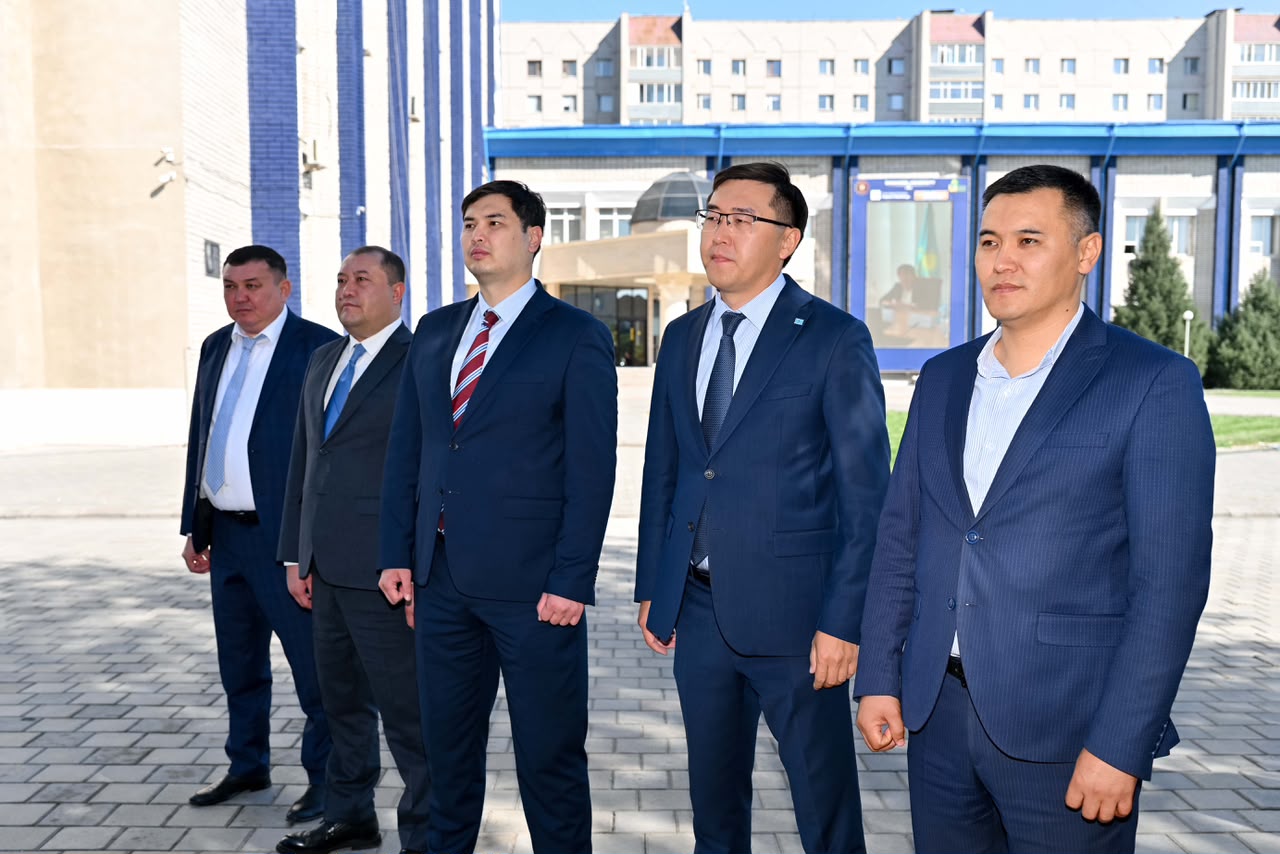 Көк костюм киген бес ер адамның ғимарат алдында тұрған суреті.