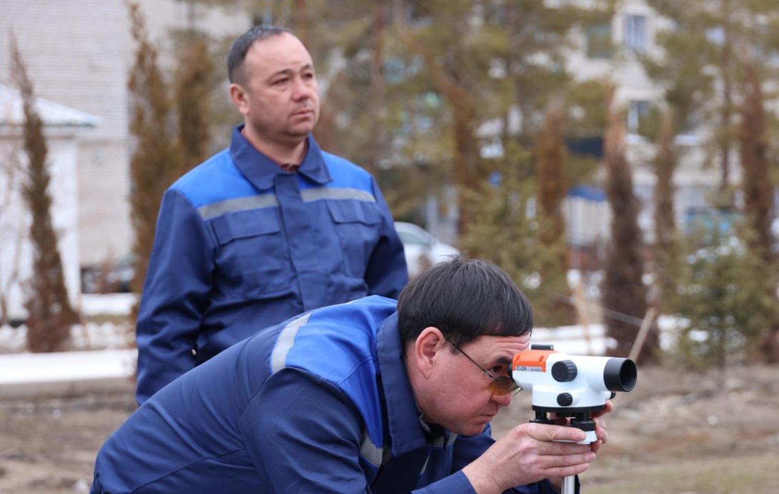 Көк киімді екі маманның сыртқы жерде геодезиялық құралмен өлшеу жүргізіп жатқаны.