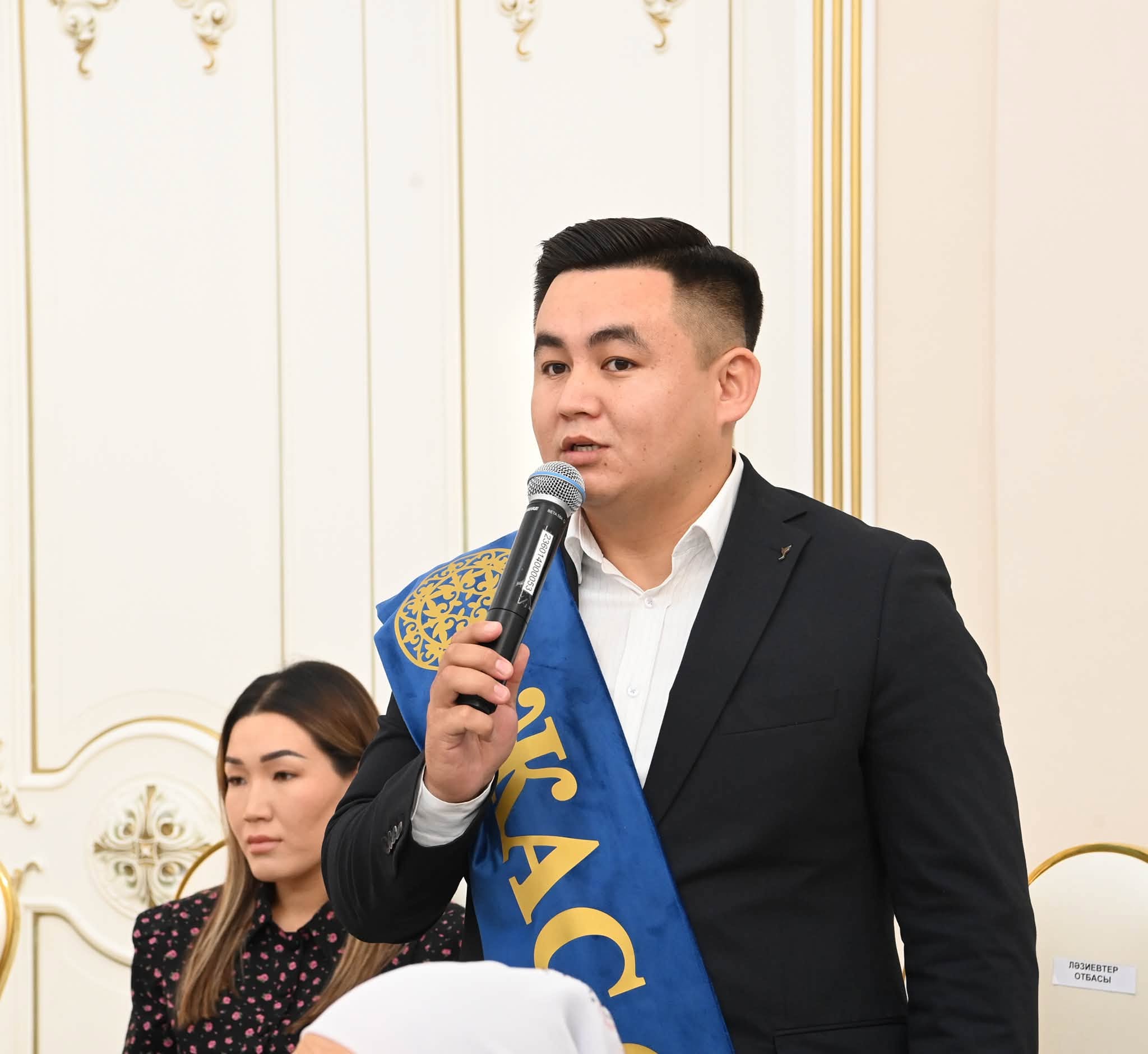 Микрофонмен сөйлеп тұрған, иығында көк жолағы бар костюм киген ер адам.