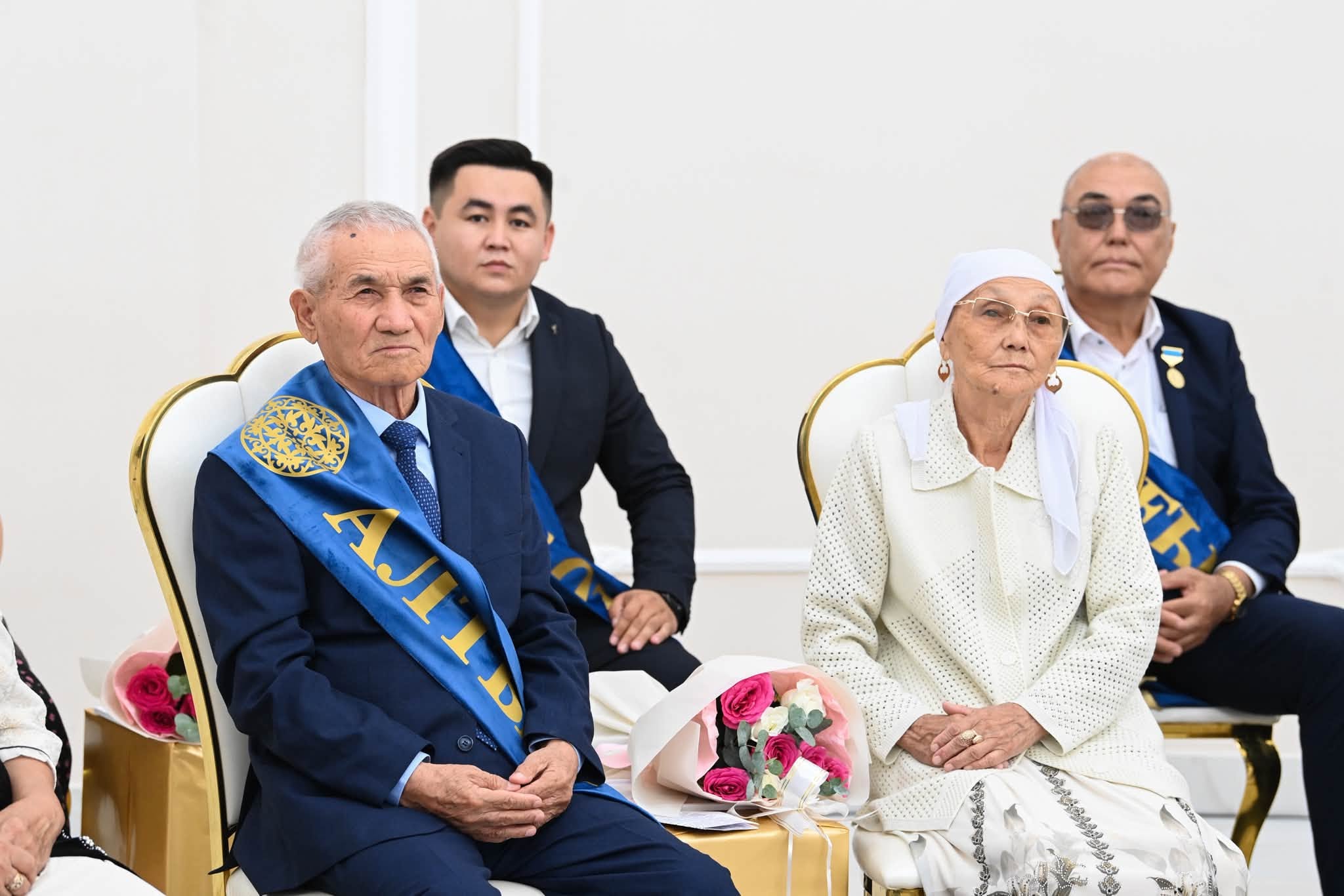 Көк лента таққан қарт ер адам мен ақ киінген қарт әйелдің салтанатты жиында отырған сәті.