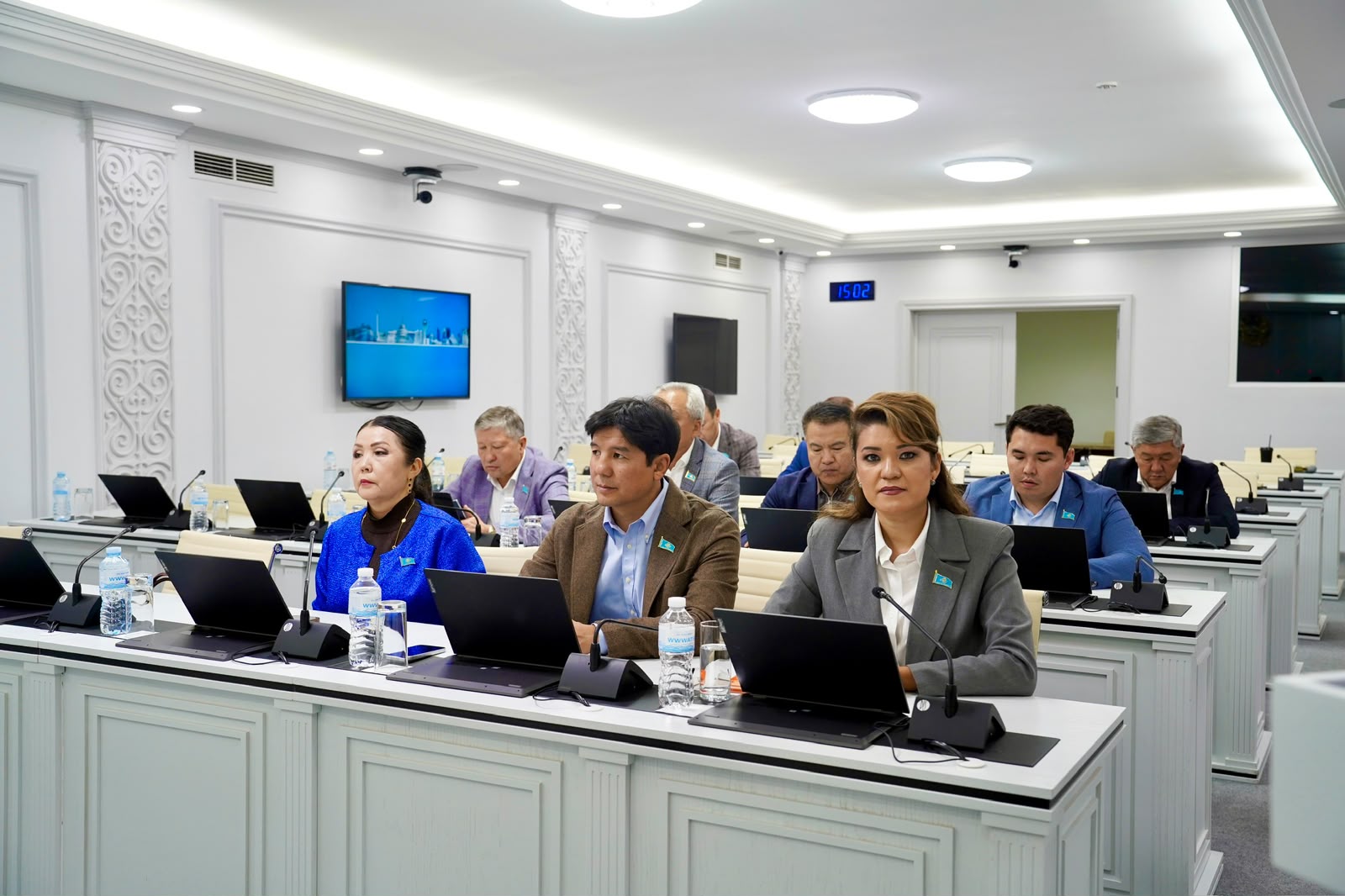 Жиналыс залында ноутбуктерімен отырған ресми киінген адамдар тобы.