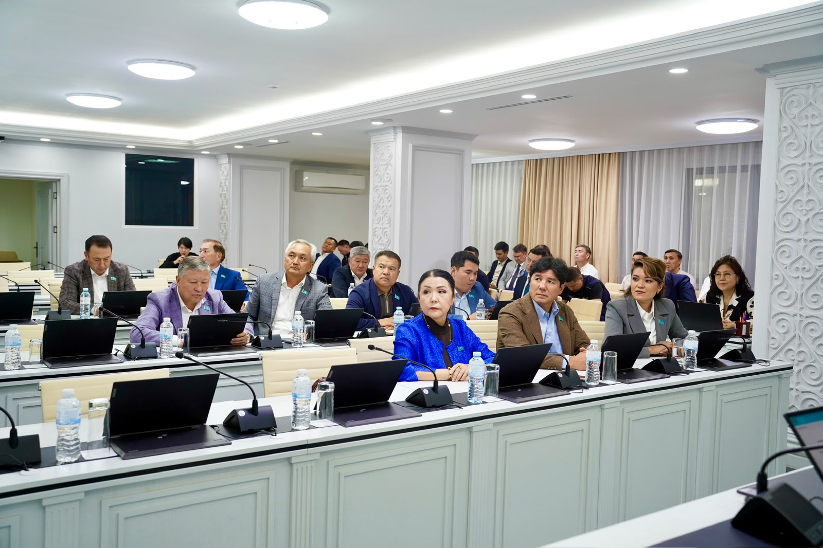 Жиналыс залында ноутбуктерімен отырған ресми киінген адамдар тобы.