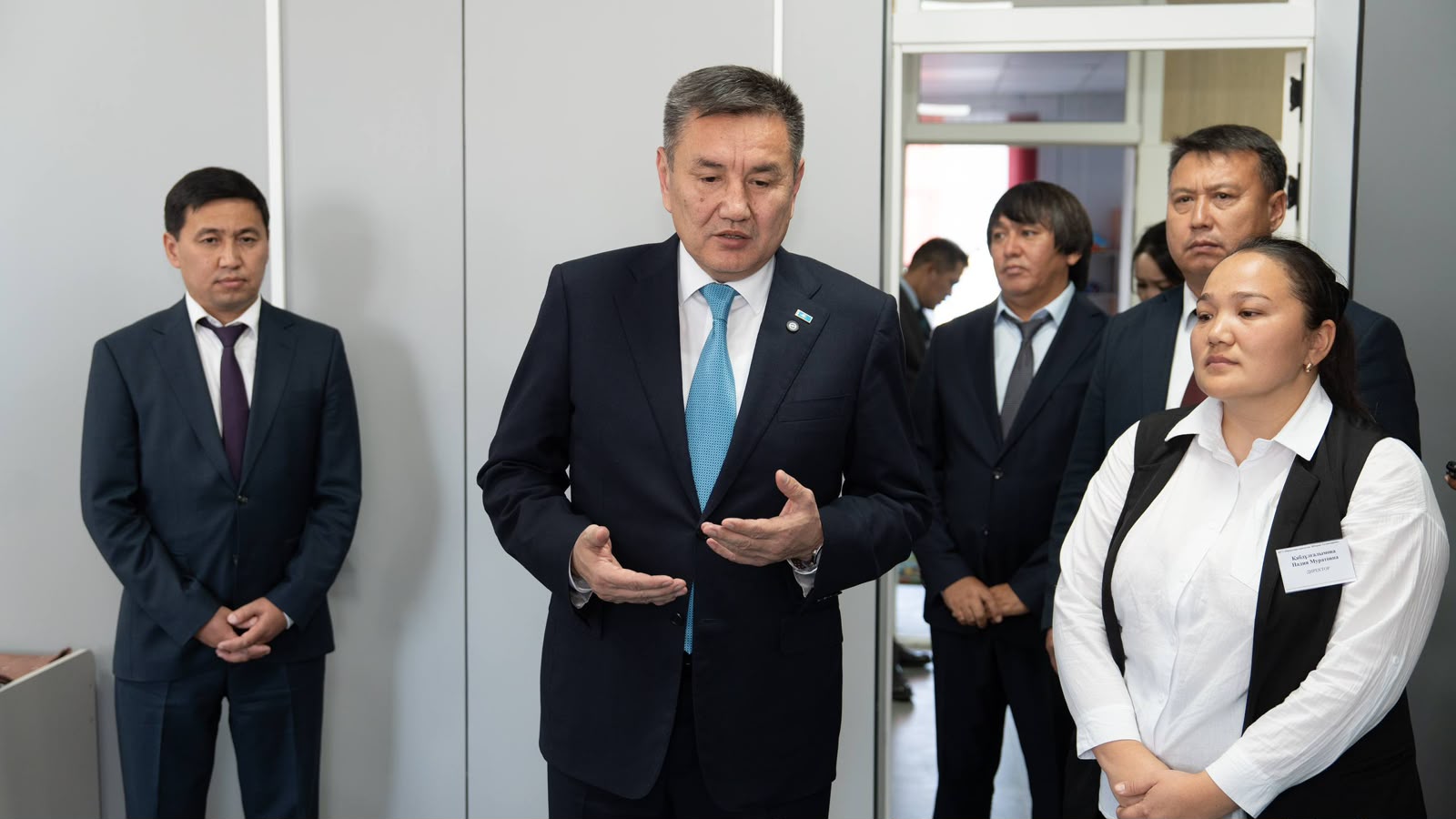 Көк галстук киген костюмді ер адамның әріптестерінің алдында сөйлеп тұрғаны.