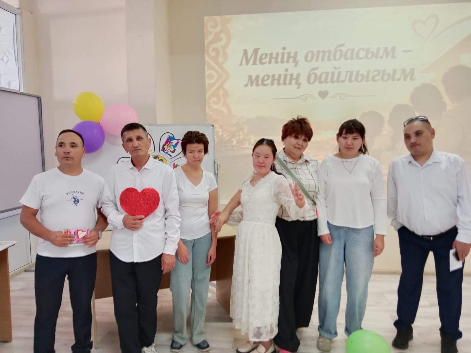 Группа людей позирует на фоне баннера с надписью на киргизском языке. Один из мужчин держит красное сердце.