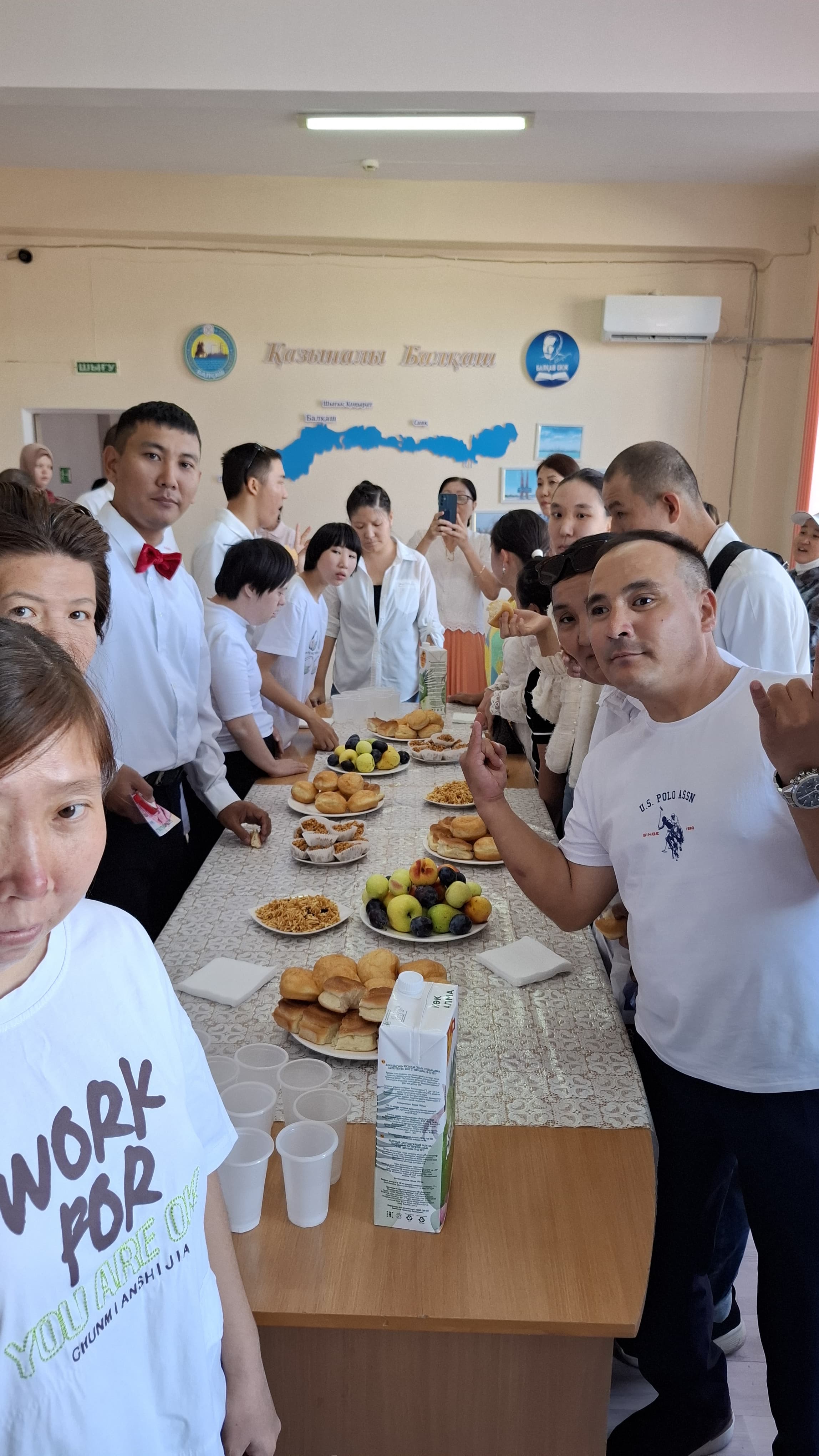 Группа людей за столом с выпечкой и фруктами в светлом помещении.