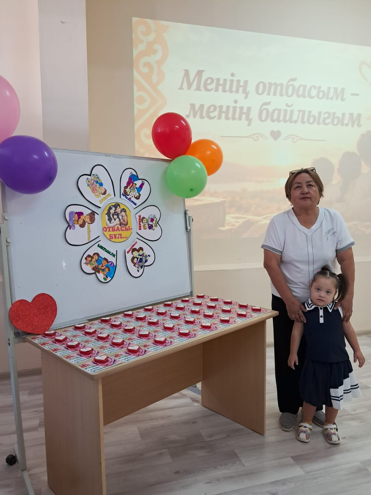 Женщина и девочка стоят рядом с праздничным столом с угощениями и доской с коллажем из семейных фото.