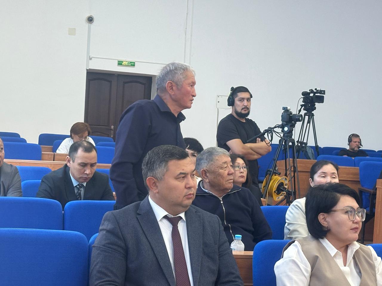 Жиналыс залындағы адамдар: тұрып сөйлеп жатқан ер адам және тыңдап отырған қатысушылар.