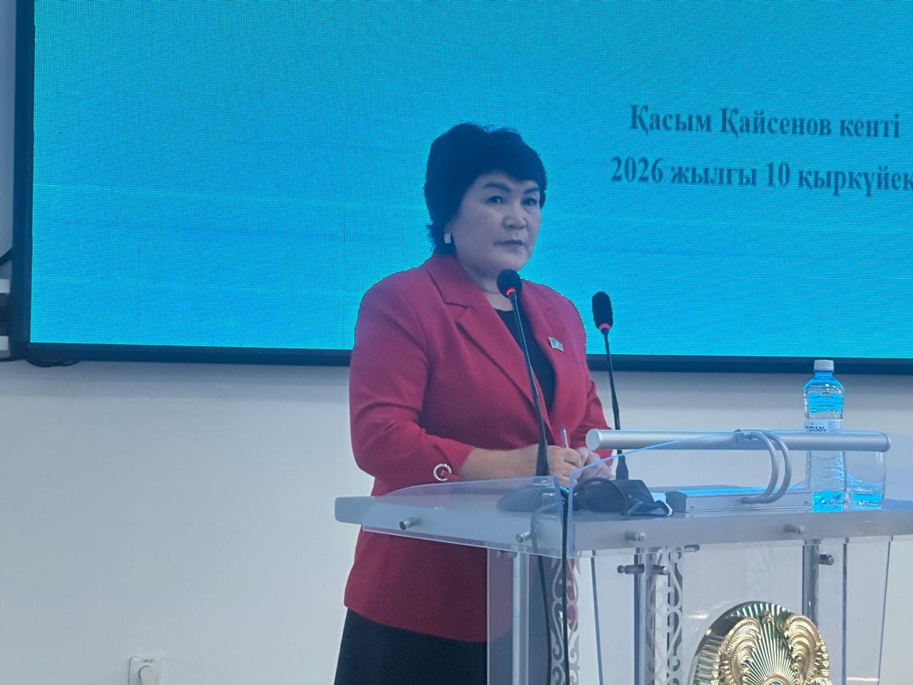 Қызыл пиджак киген әйел адам трибунада сөйлеп тұр. Артында көк экранда мәтін жазылған.