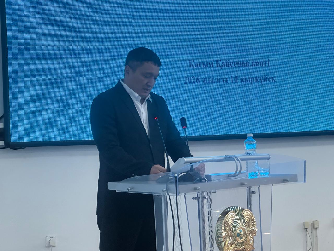 Костюм киген ер адам трибунада тұрып, артындағы көк экранда мәтін жазылған жерде сөйлеп тұр.