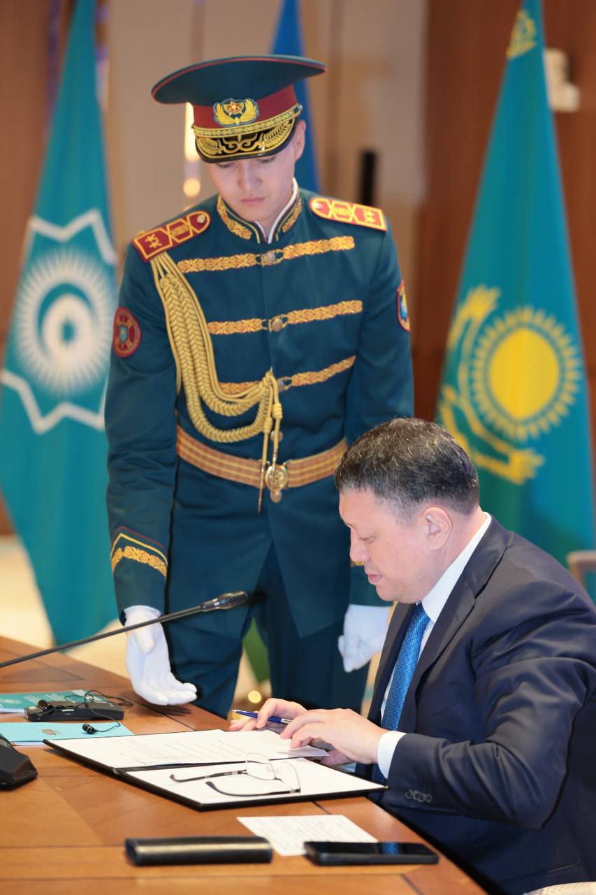 Қазақстан туларының алдында құжатқа қол қойып жатқан Ержан Саденов және оның жанында тұрған салтанатты киімдегі күзетші.