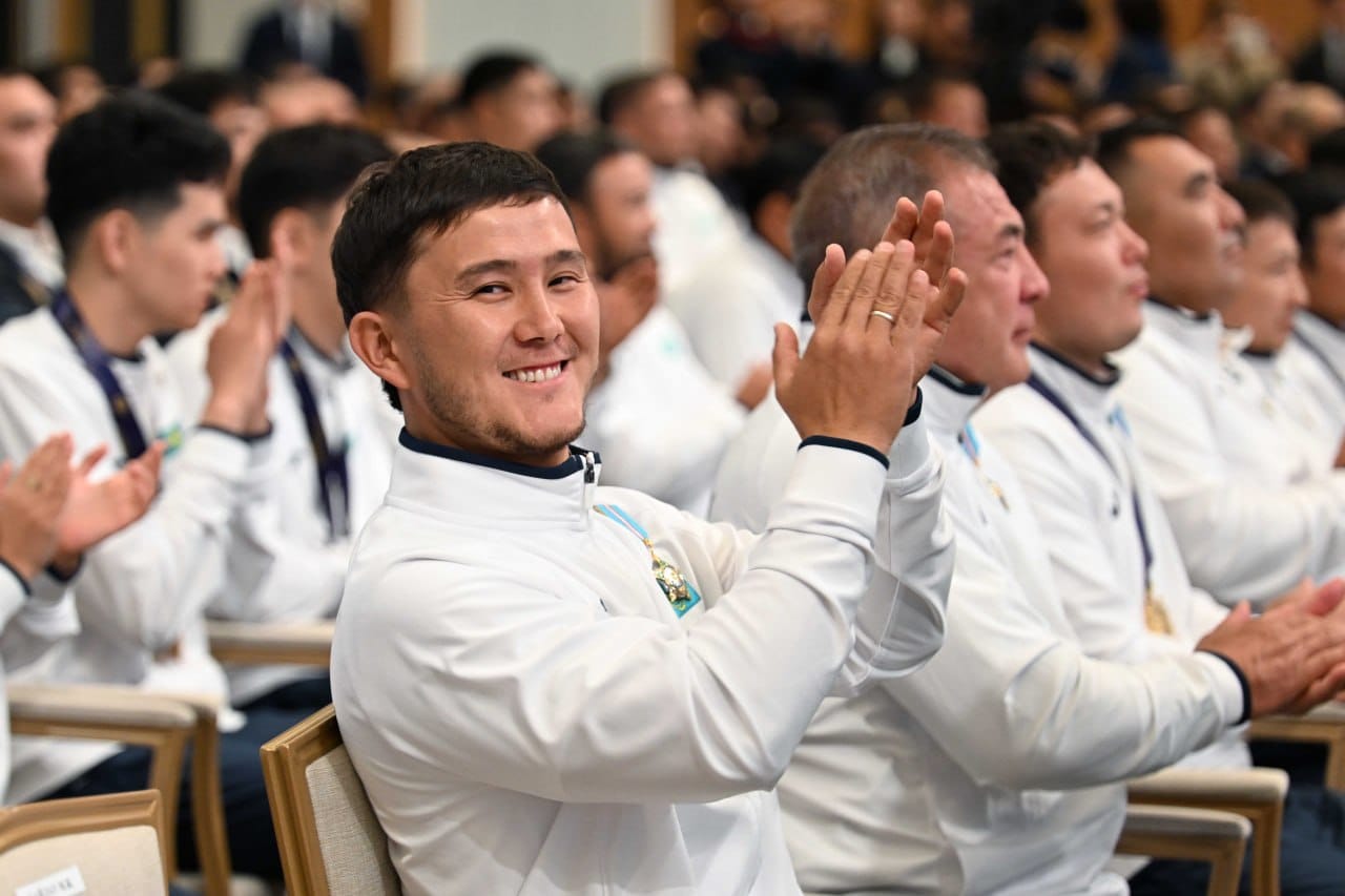 Ақ спорттық киім киген, қол шапалақтап тұрған күлімсіреген ер адам.