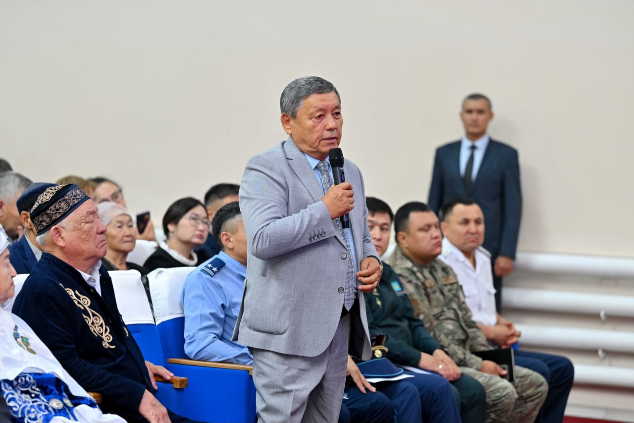 Сұрақ қойып жатқан сұр костюмді ер адам және аудиториядағы тыңдаушылар.