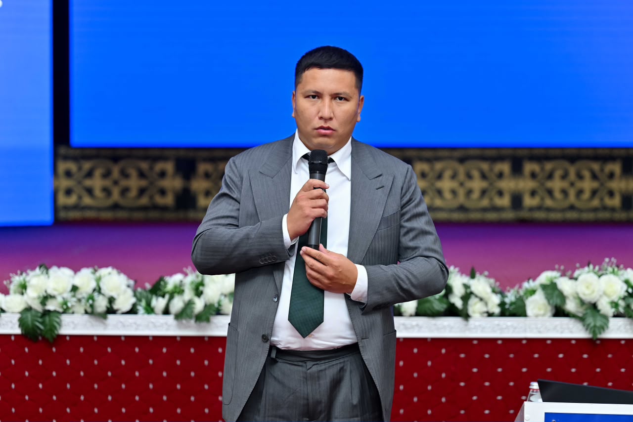 Сұр костюм киген ер адам микрофонмен сөйлеп тұр.