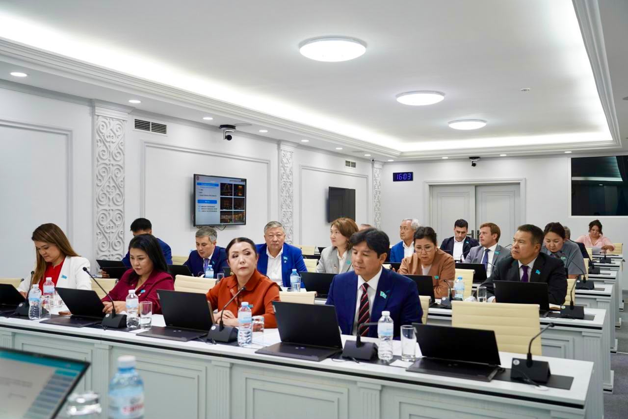 Жиналыс залында ноутбуктермен жұмыс істеп отырған ресми киінген адамдар тобы.