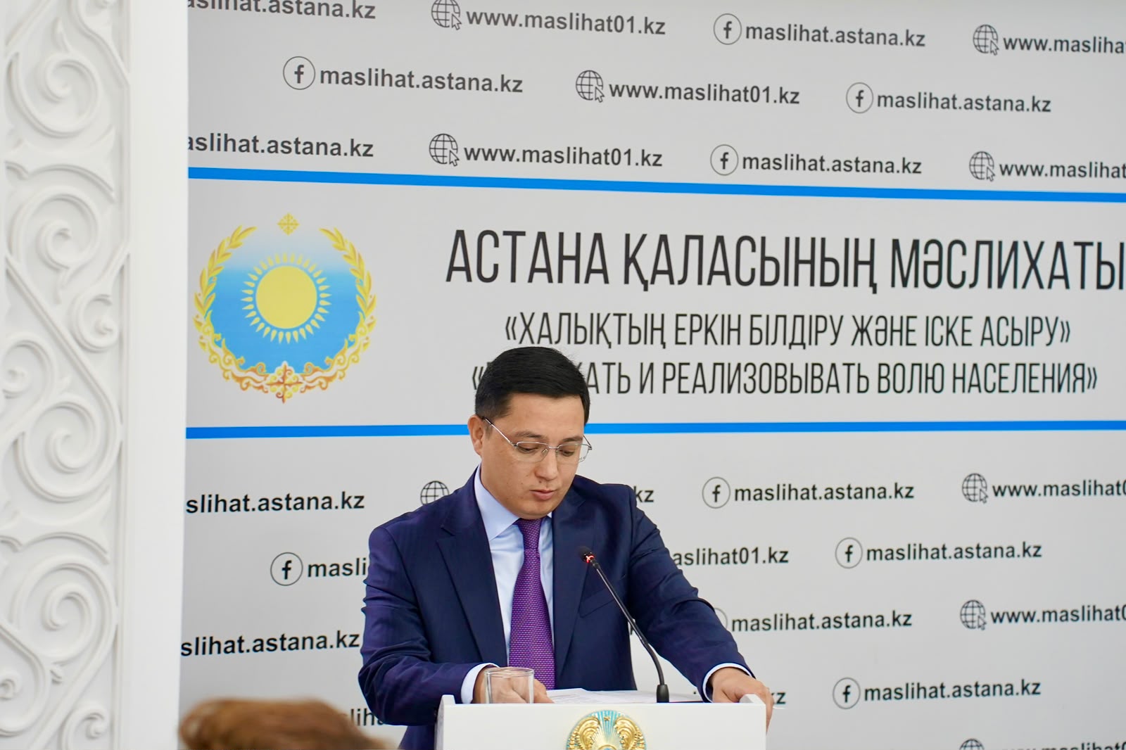 Астана қаласының мәслихатының алдында сөйлеп тұрған костюм киген ер адам.