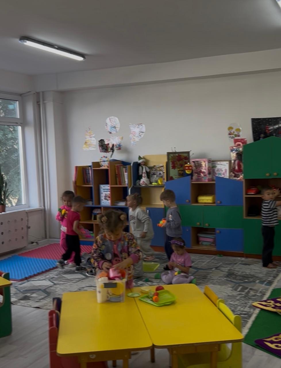 Группа детей играет в светлой игровой комнате детского сада с разноцветной мебелью и игрушками.