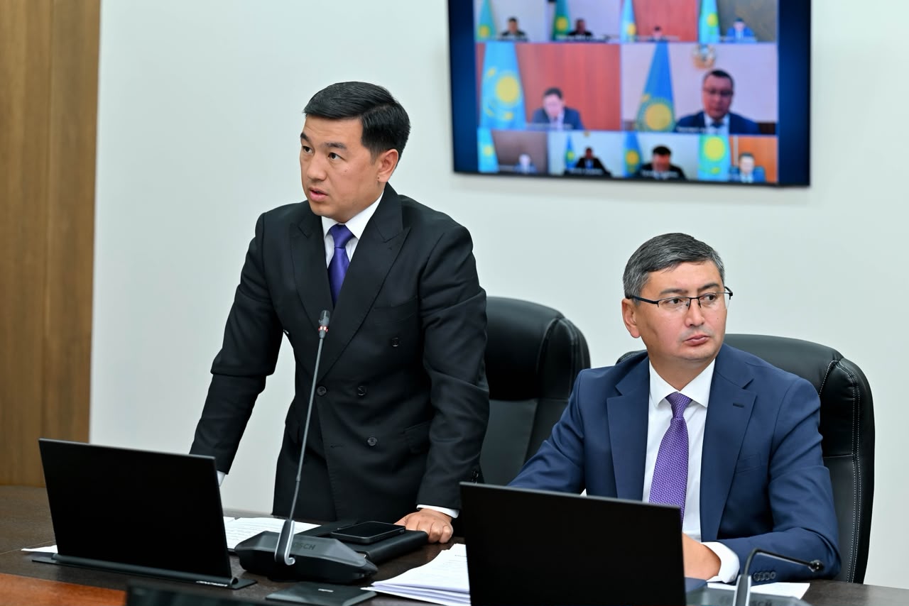 Костюм киген екі ер адам жиналыста, артқы жақта бейнеқоңырау экраны көрінеді.