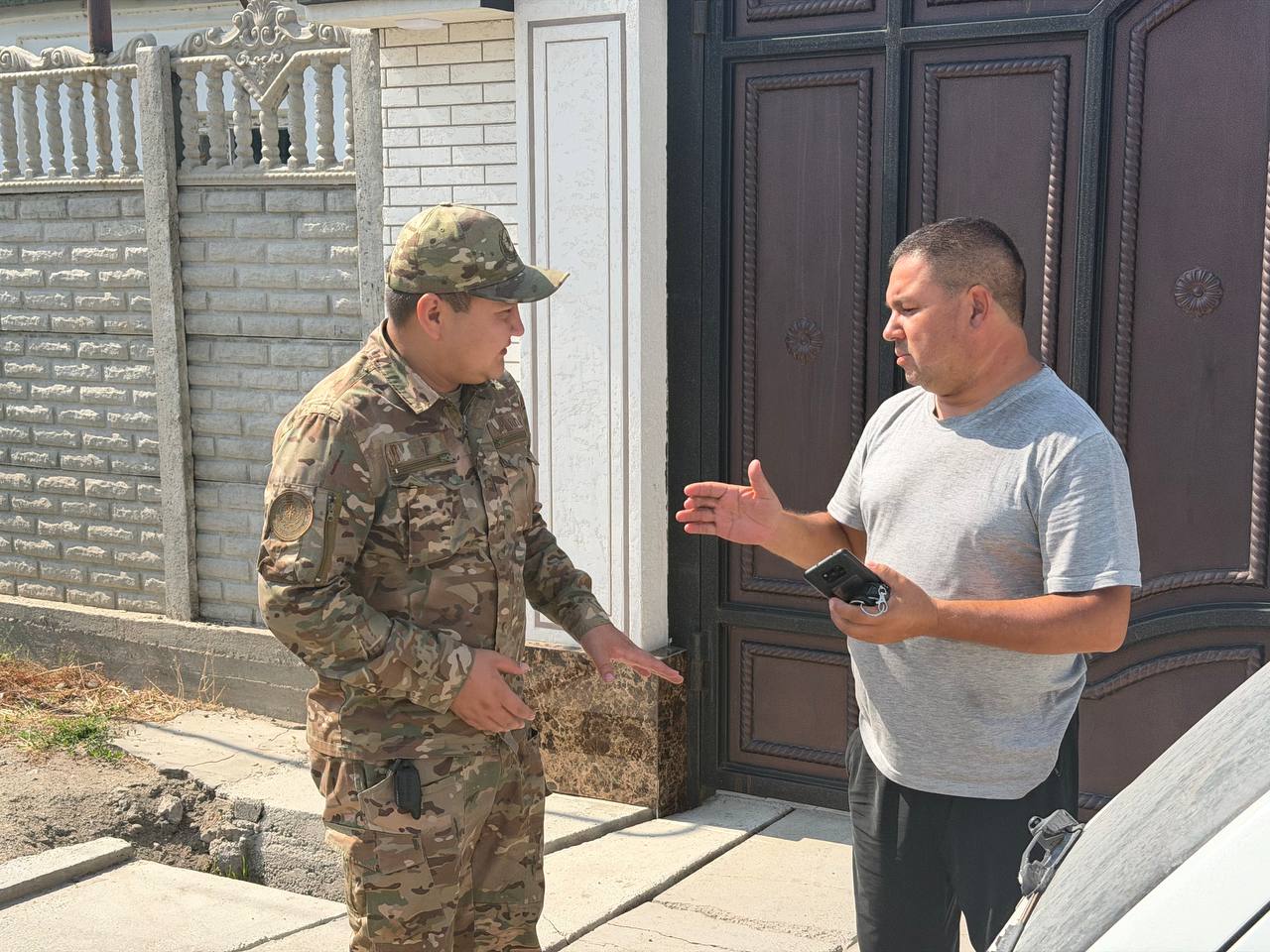 Әскери формадағы адам мен қарапайым киінген ер адамның үйдің алдында сөйлесіп тұрғаны.